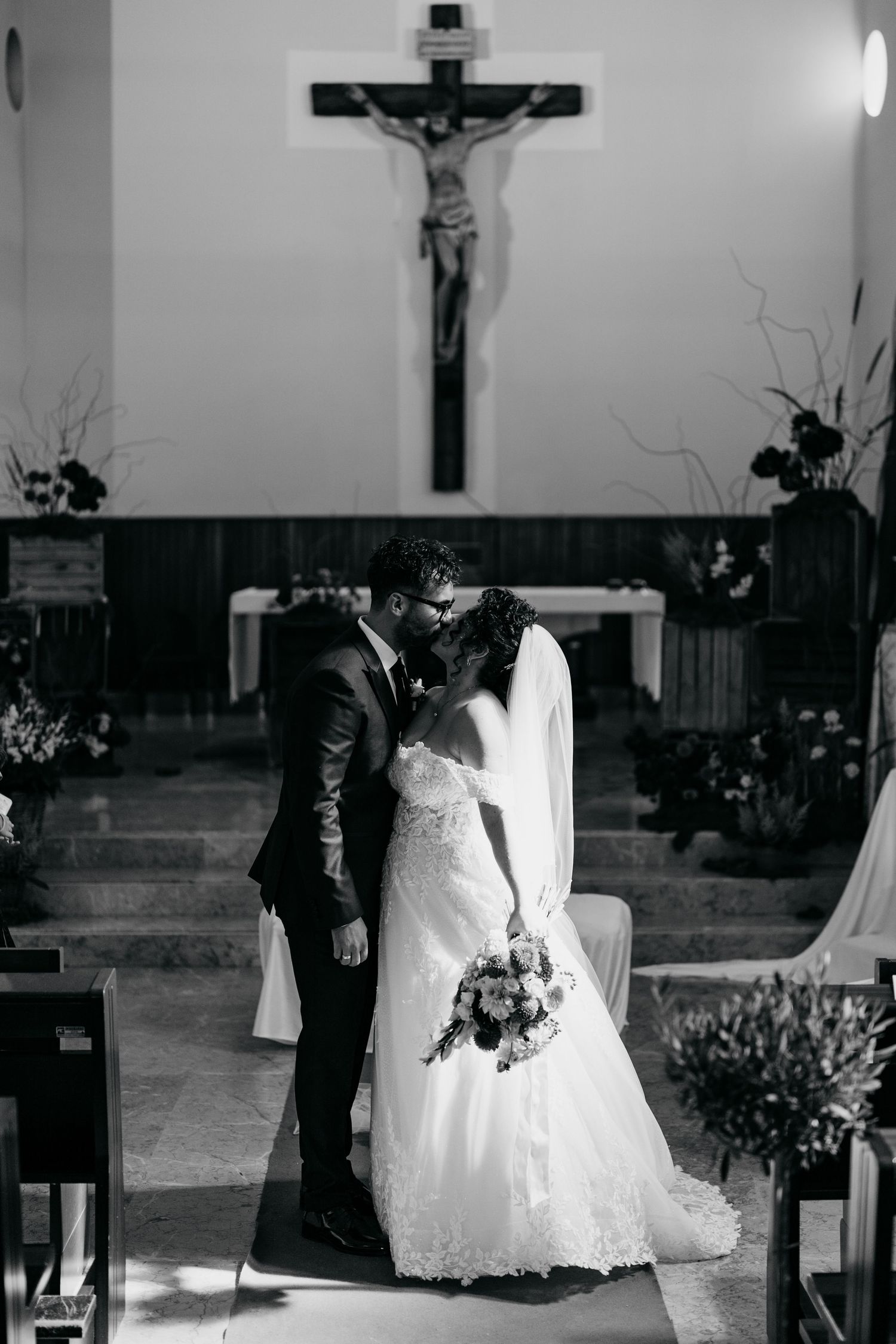 Bride and groom sharing a kiss at the altar of a church with a crucifix in black and white.
