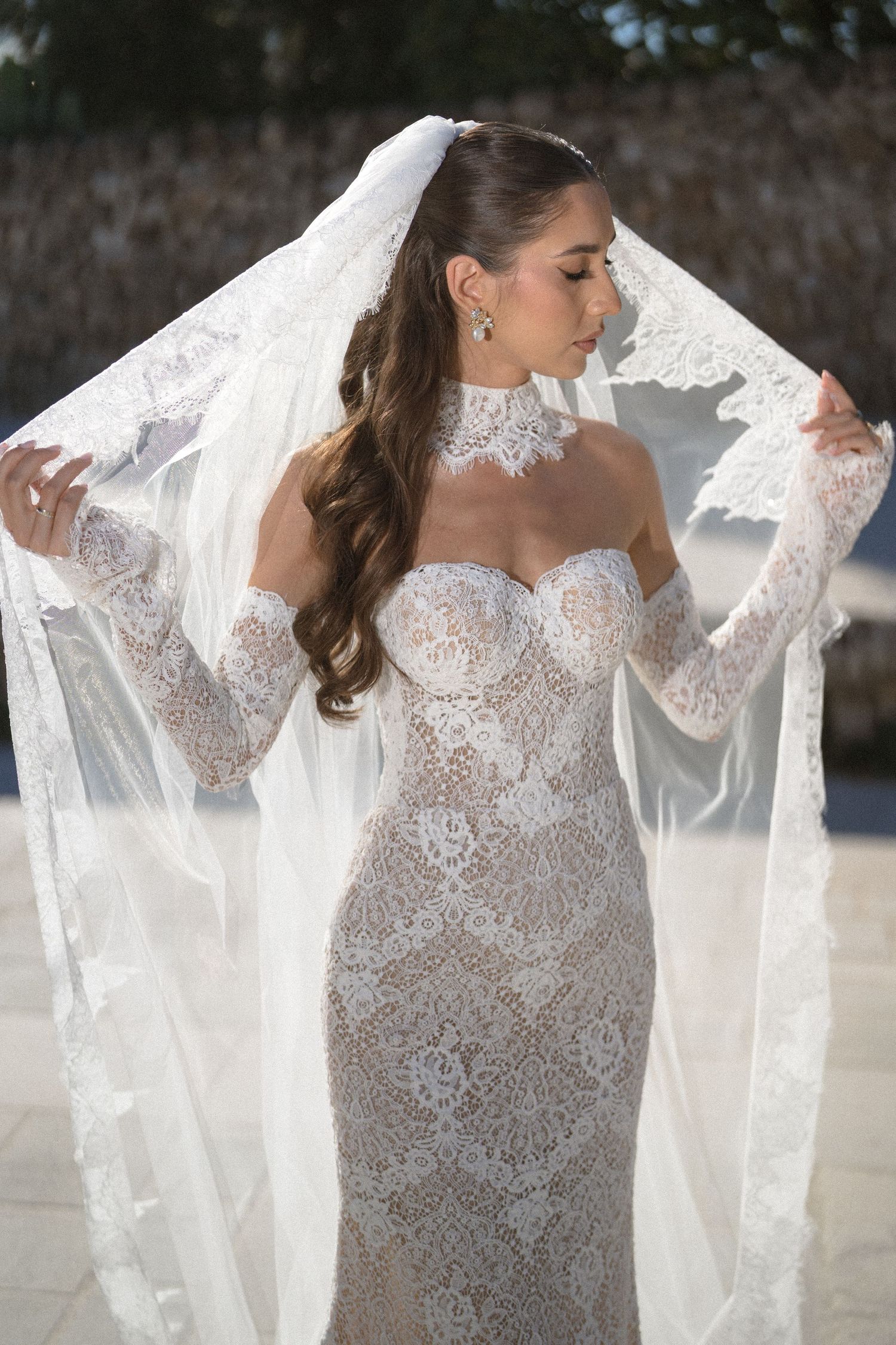 Bride in a strapless lace wedding gown with high collar, sheer long sleeves and a flowing veil, hair styled in loose waves.
