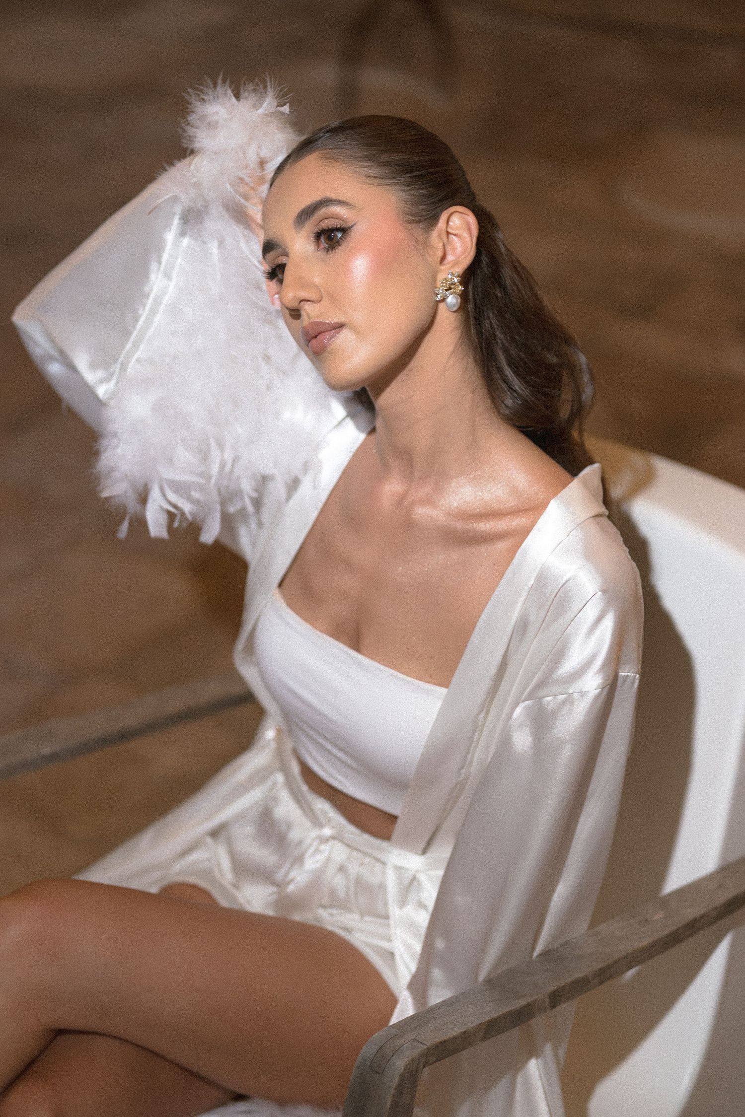 A woman in a white satin robe and crop top sits in a chair, leaning back with a feathered sleeve raised beside her head.