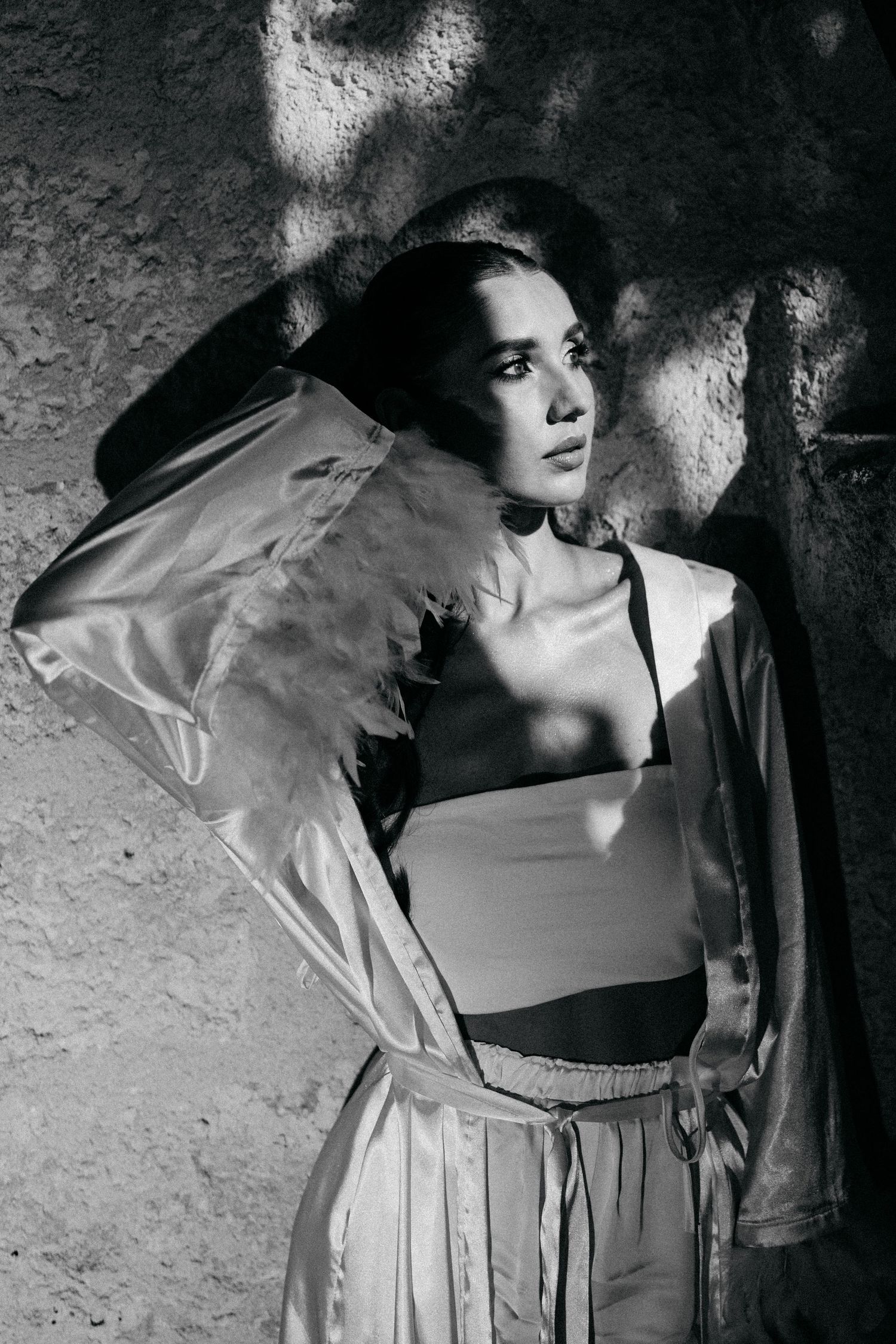 Black and white portrait of a woman in a satin robe with feather-trimmed sleeve posing against a textured wall.