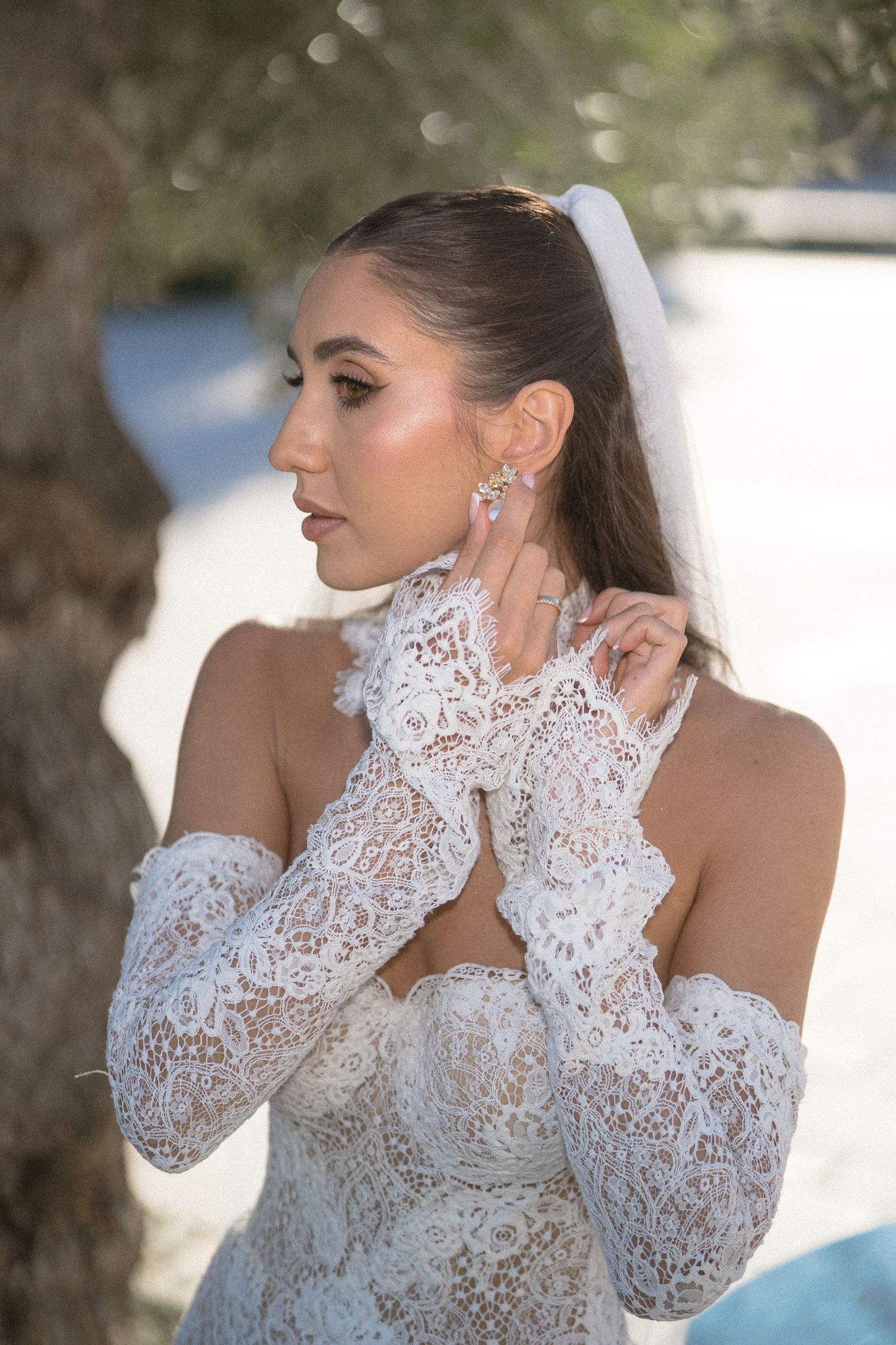 Bride in profile touching her earring, wearing a lace off-shoulder gown with long lace sleeves under an olive tree.