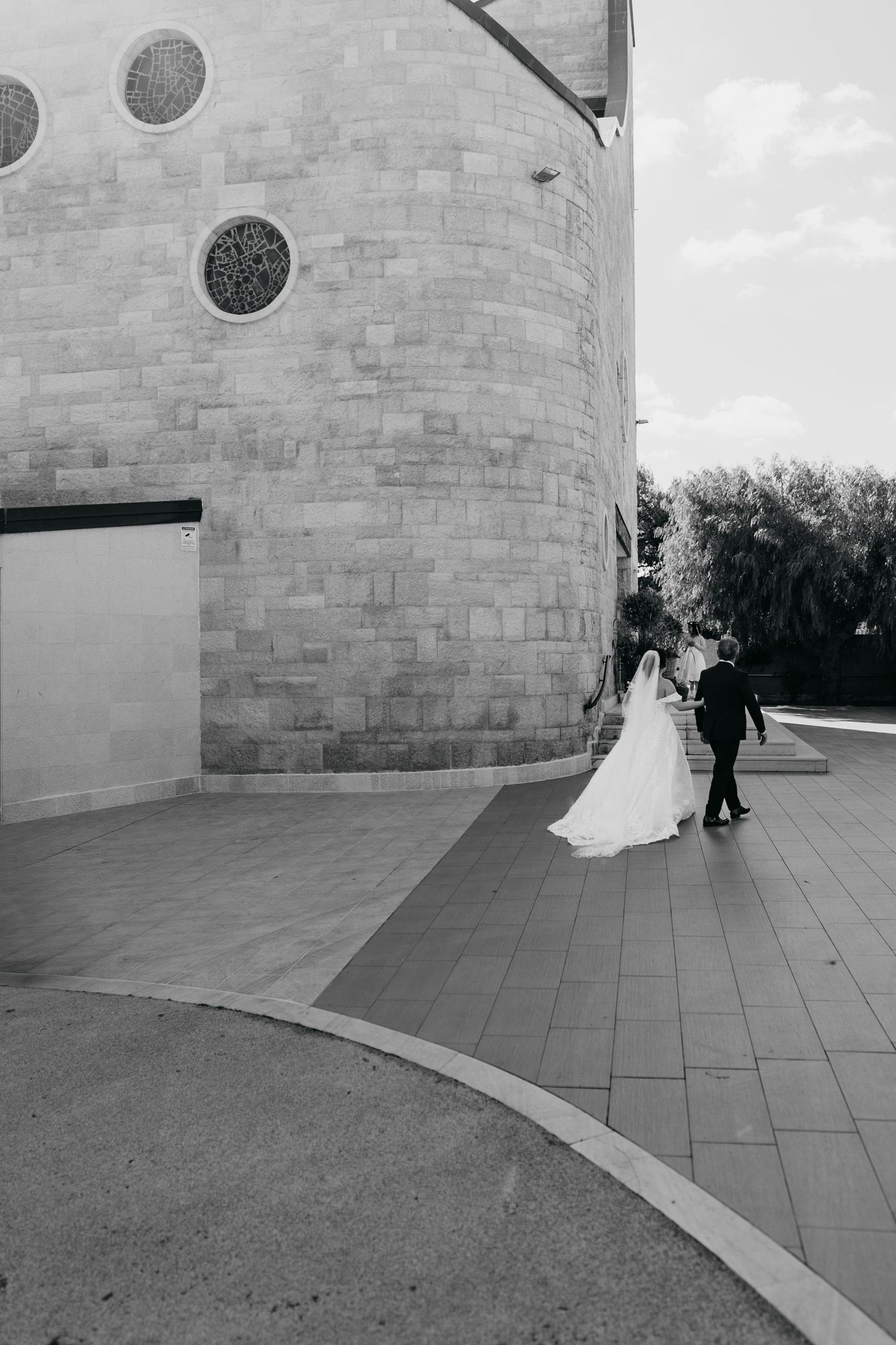 Black and white photo of bride and groom walking along curved stone church exterior on wedding day.