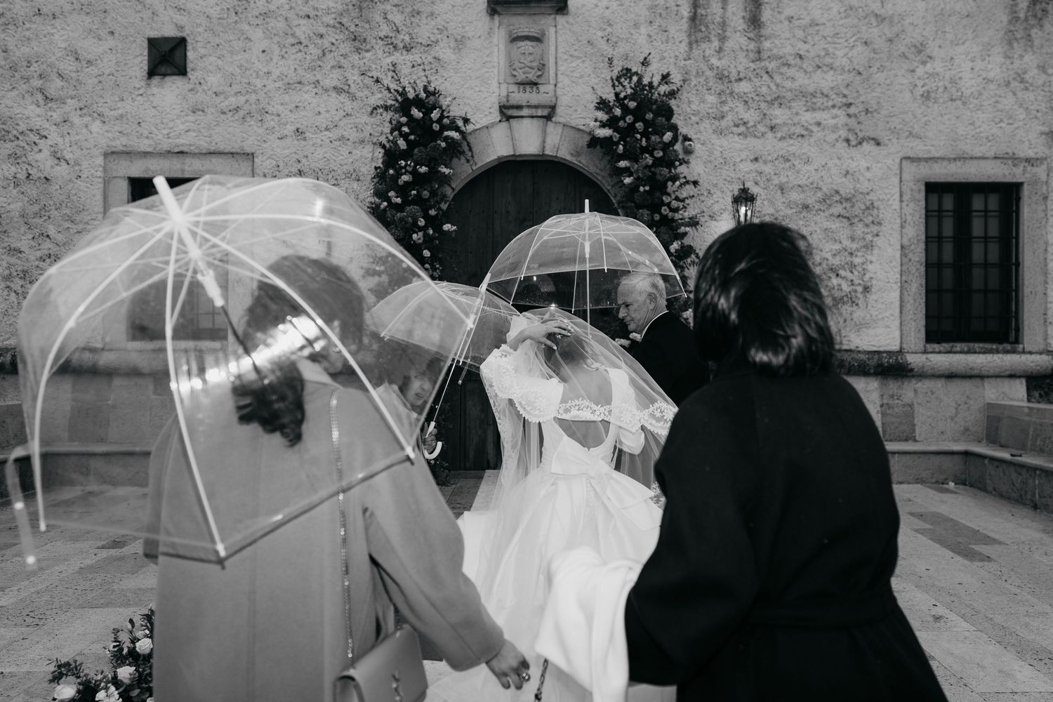 Bride with transparent umbrellas in rainy black and white wedding photo outside historic venue.