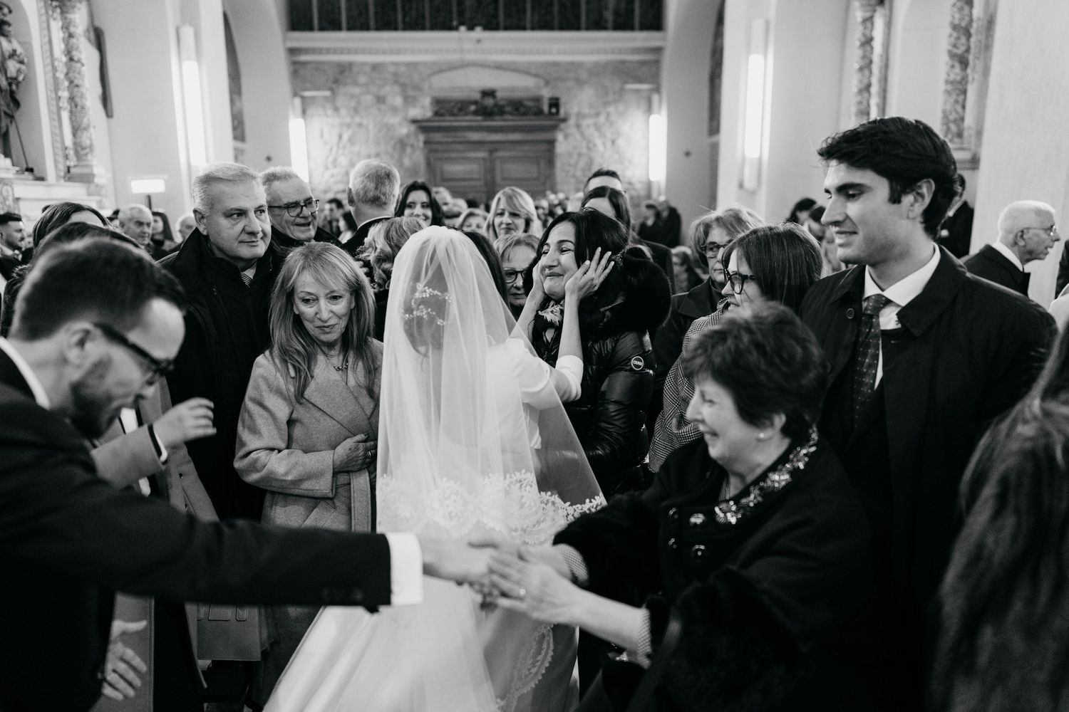 A bride in a veil walks down the church aisle surrounded by smiling wedding guests in a black and white photo.