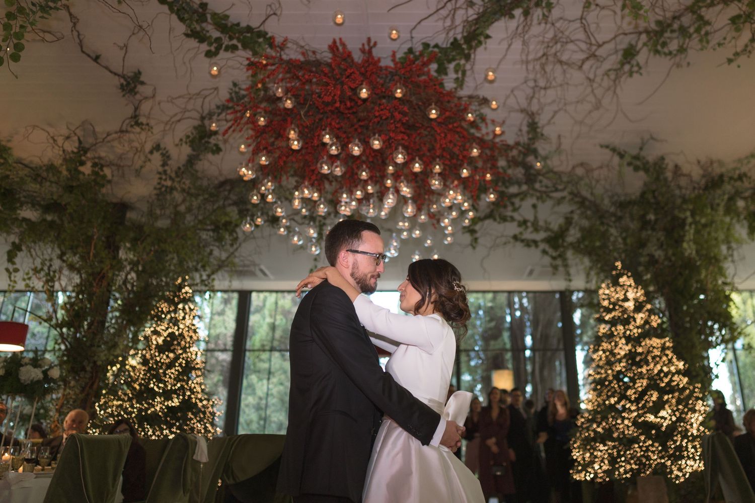 Bride and groom share first dance under stunning floral chandelier surrounded by decorated Christmas trees.