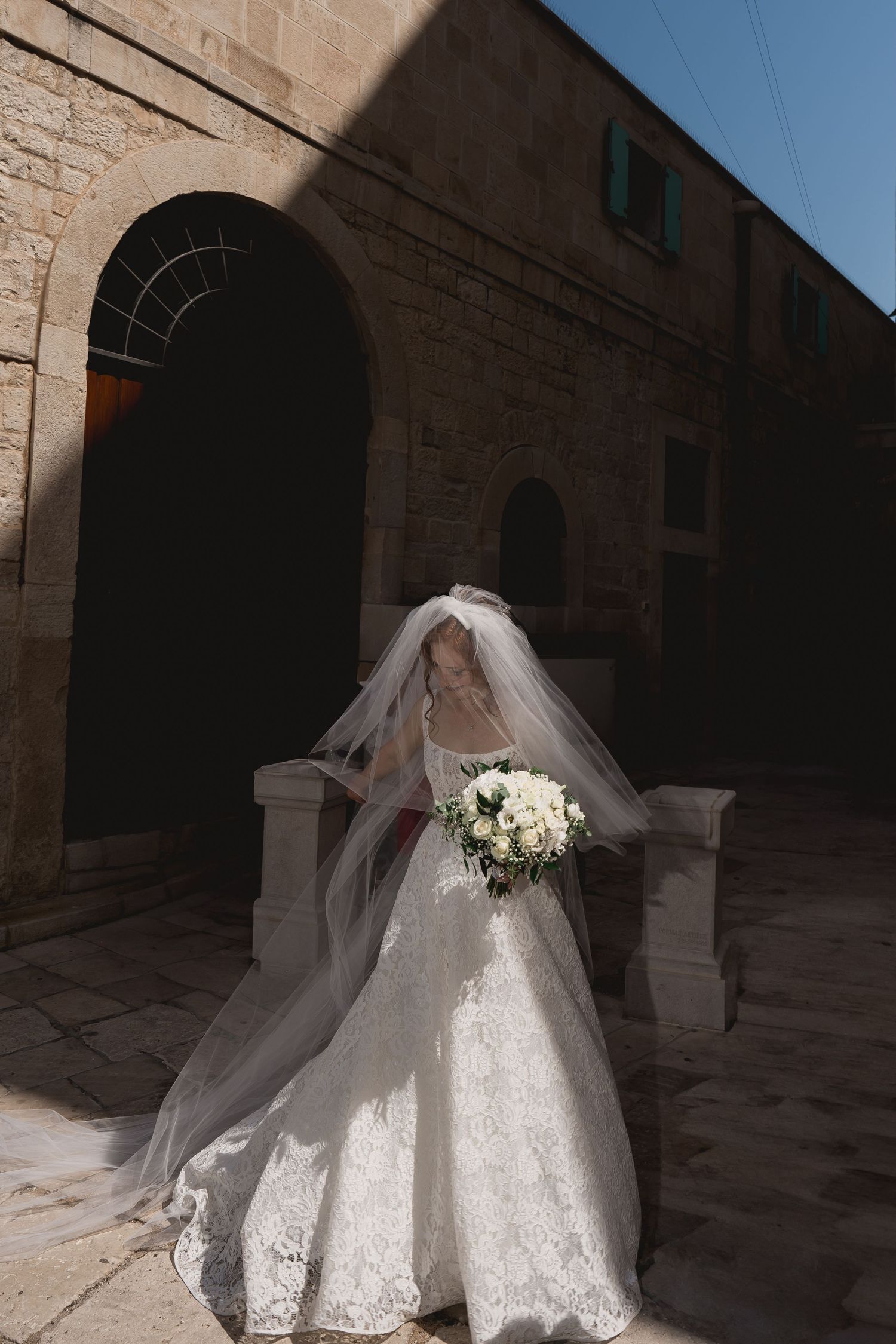 A bride in a flowing white lace wedding gown and veil poses in a stone archway corridor.