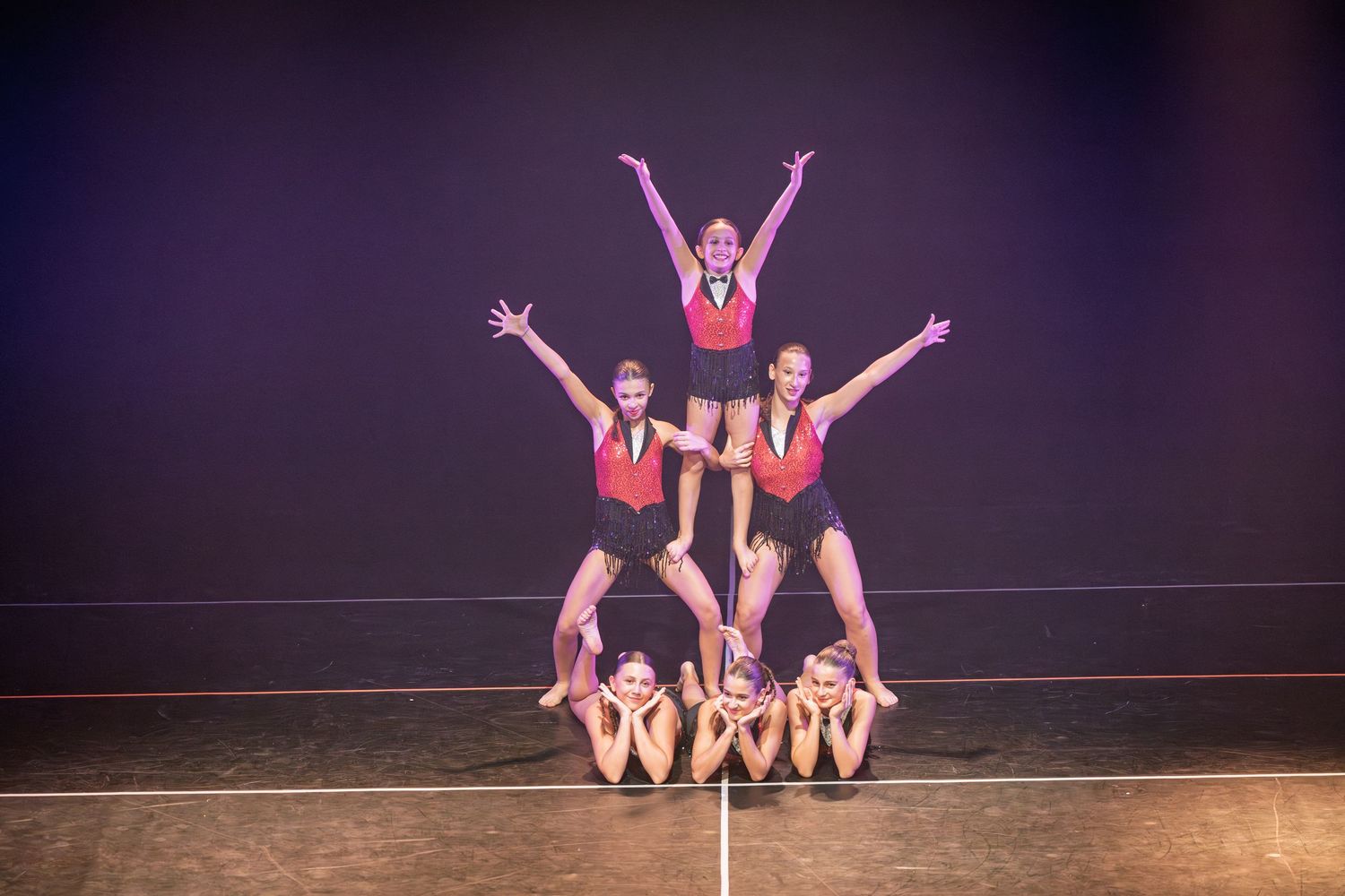 Dancers in pink costumes perform a dynamic group routine on stage with dramatic poses and lifts.
