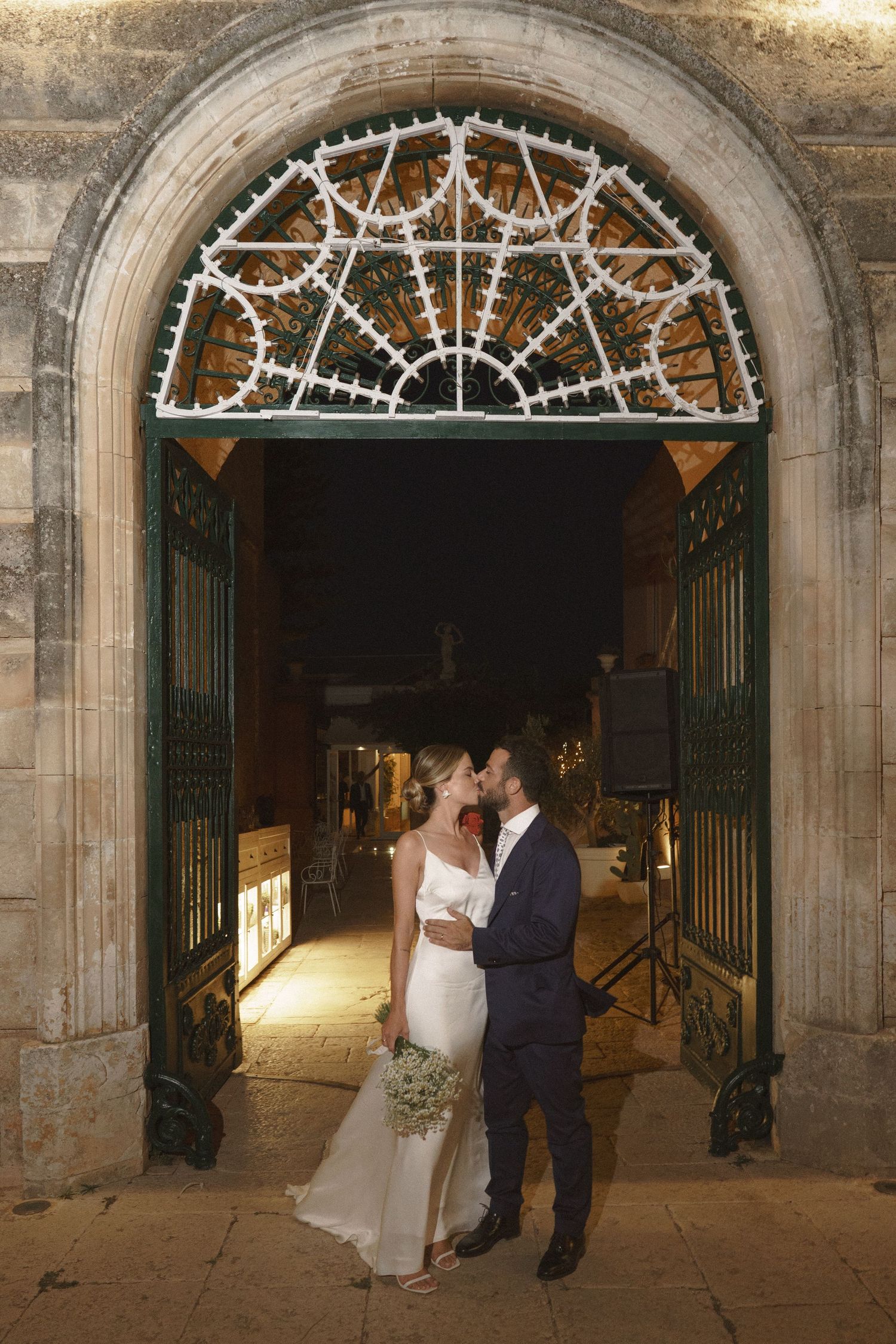 A romantic wedding moment under an ornate stone archway with decorative metalwork at night.