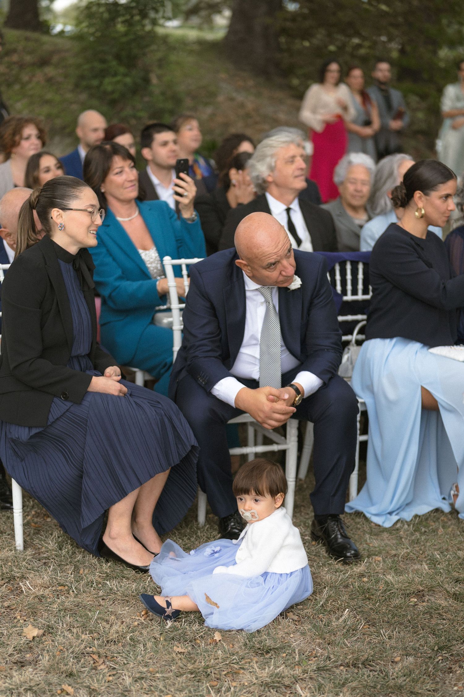 Wedding guests seated on white chairs on a grassy lawn during an outdoor ceremony.