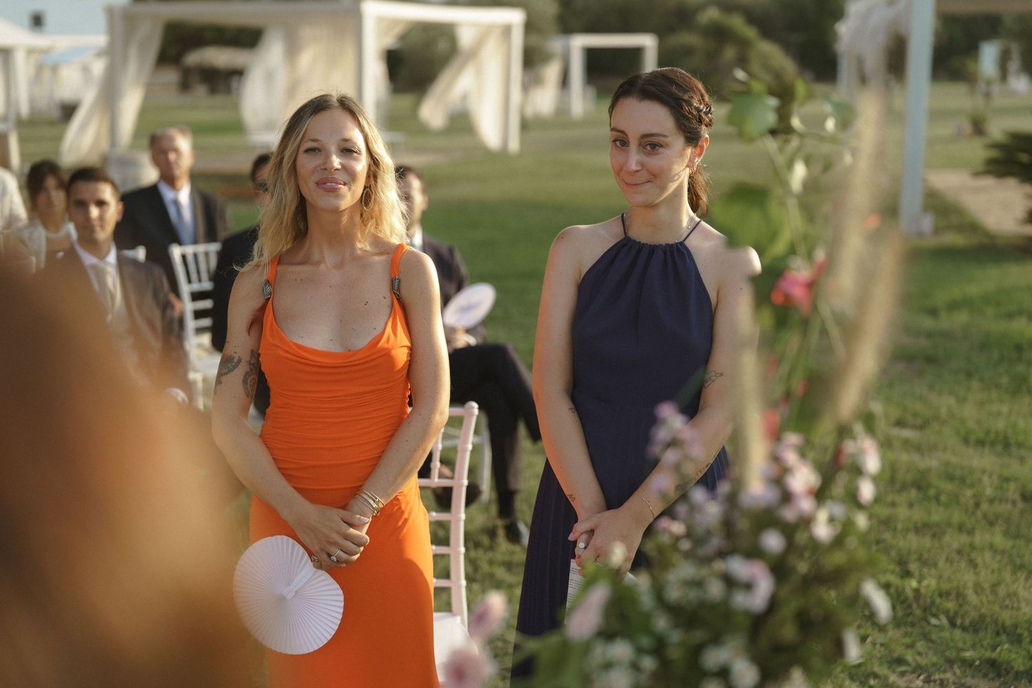 Wedding guests in colorful dresses stand along a flower-lined aisle at an outdoor ceremony.