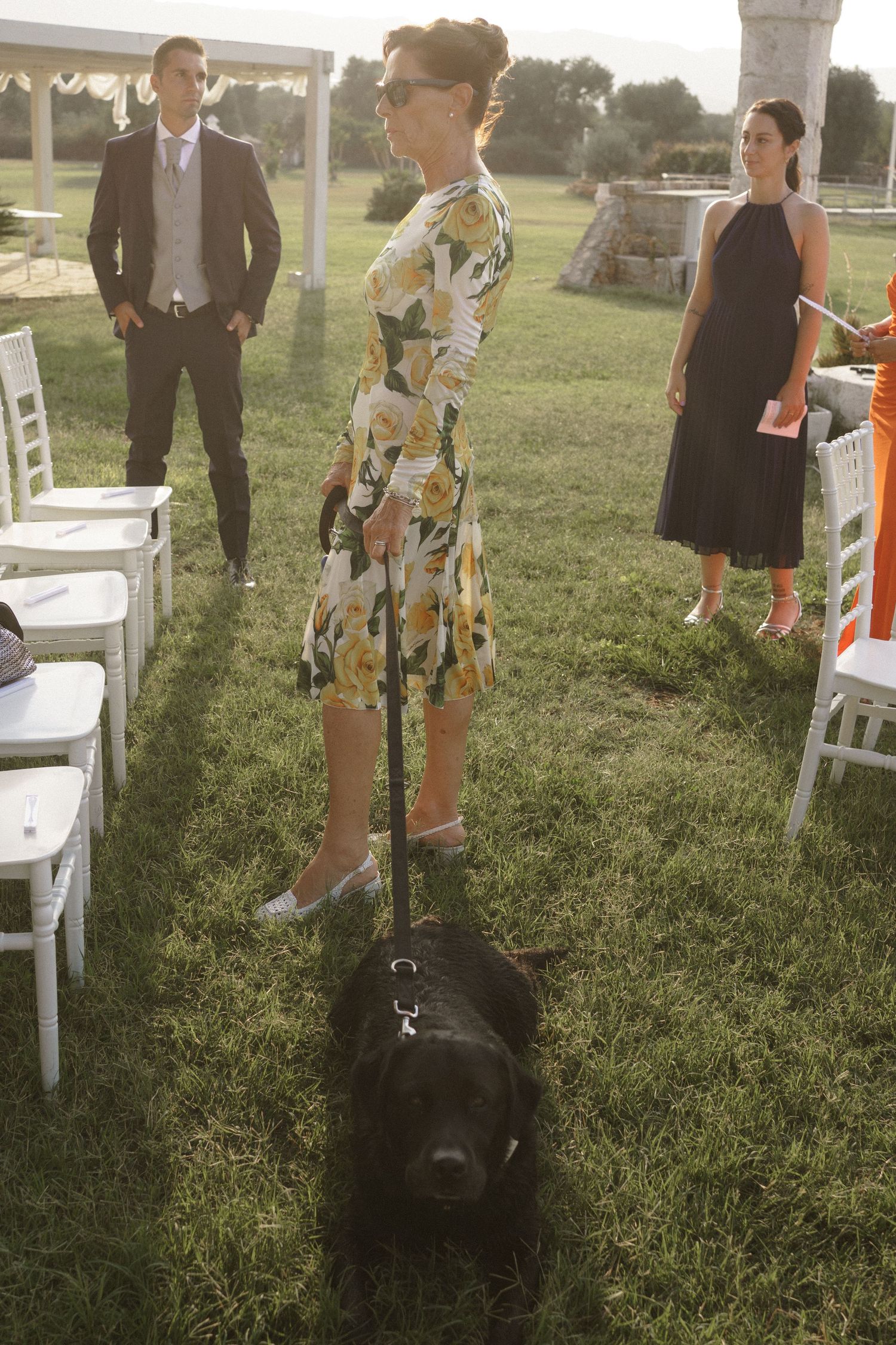 An outdoor wedding scene with white chairs arranged on grass and a person wearing a floral dress standing with a black dog.
