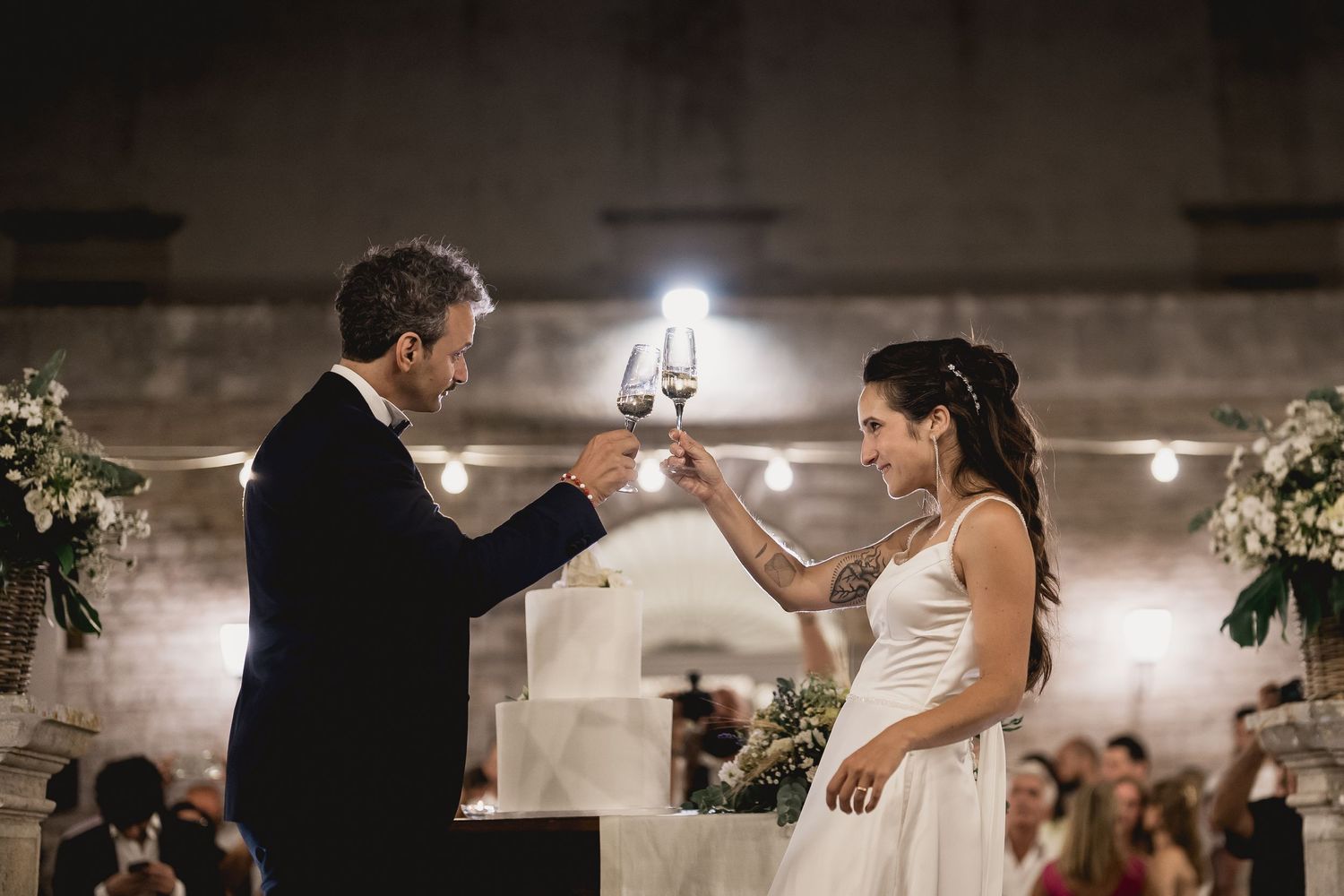 Elegant wedding couple raises champagne glasses for a toast in a beautifully decorated venue with soft lighting.