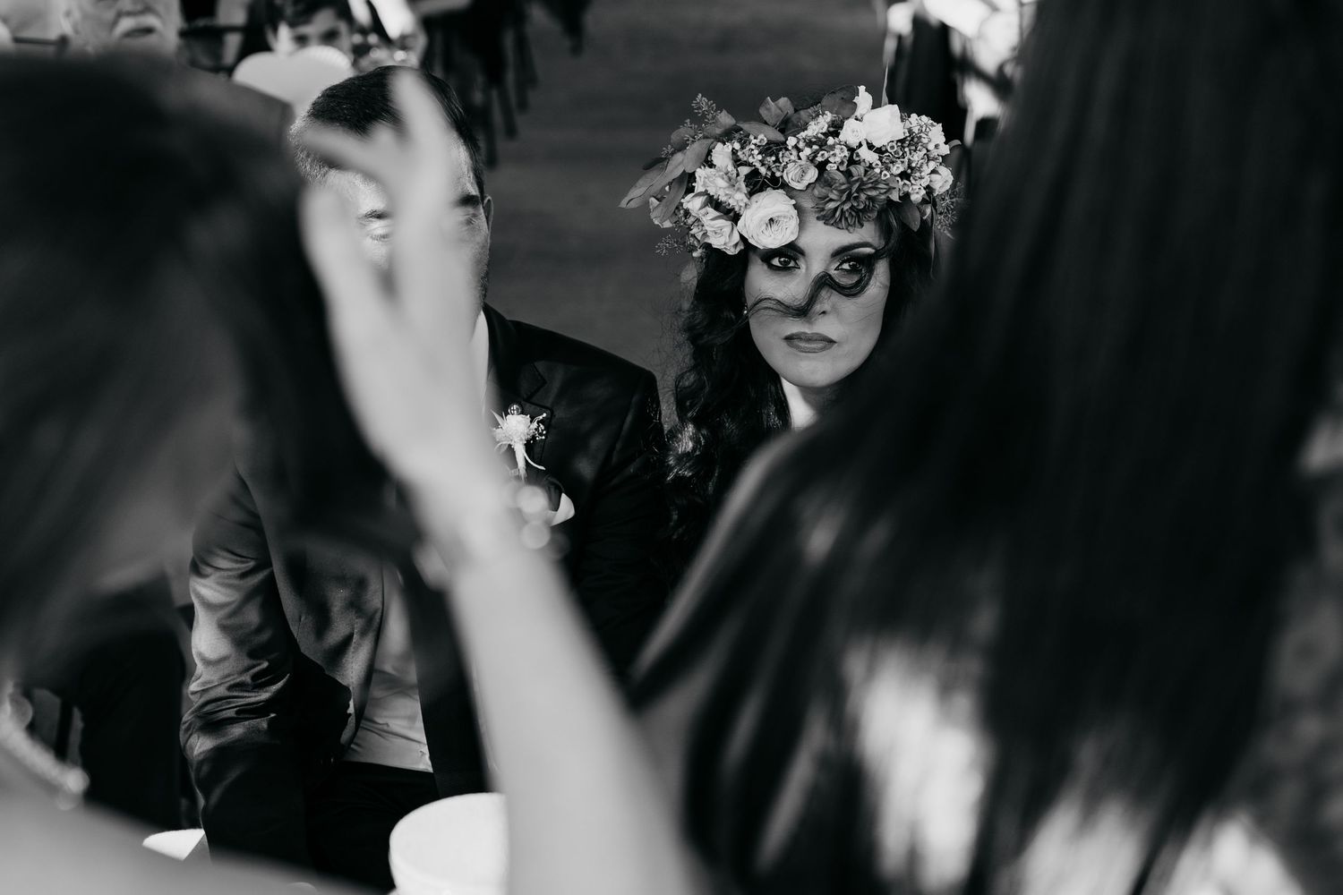 Black and white photo of someone wearing an ornate floral crown at a festive gathering.