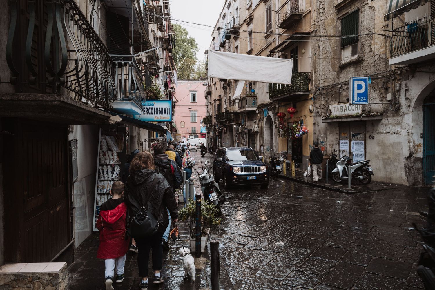 Narrow cobblestone street in Naples with laundry hanging between weathered buildings on a rainy day.