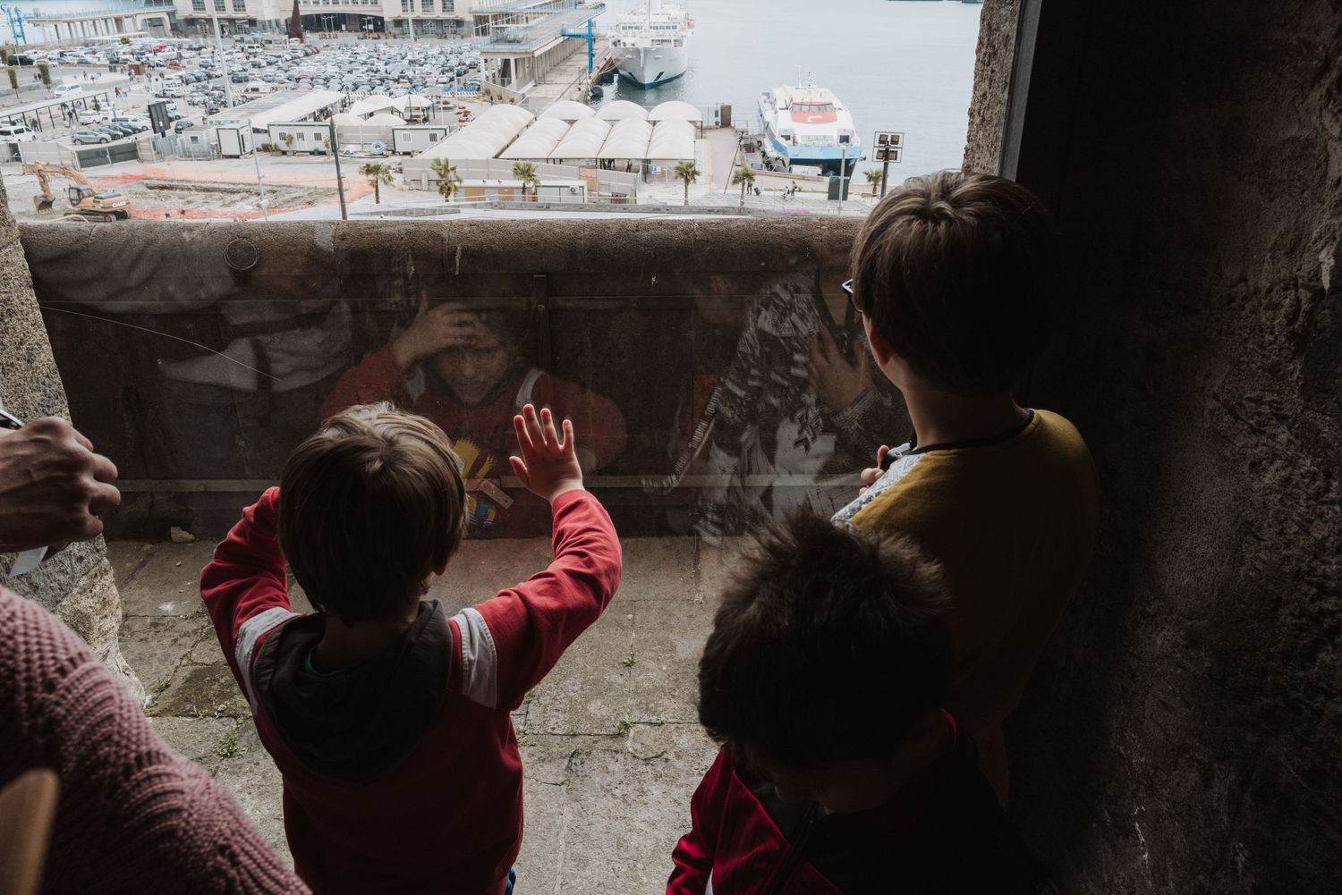 Three children look out through a window frame at an urban landscape with buildings in the distance.