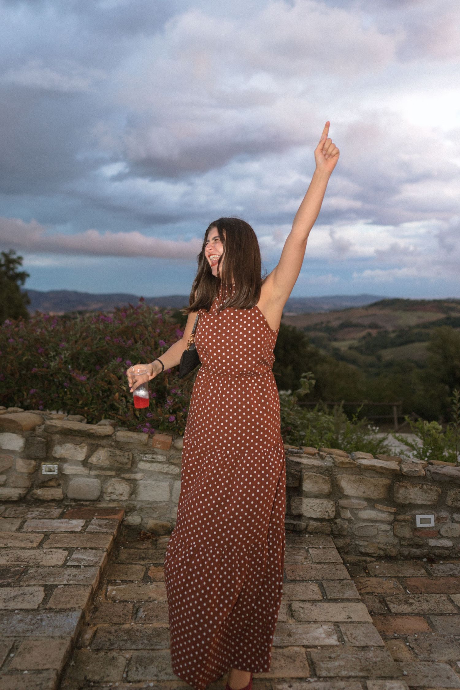 Woman in polka dot dress dancing with raised arm against cloudy sunset landscape.