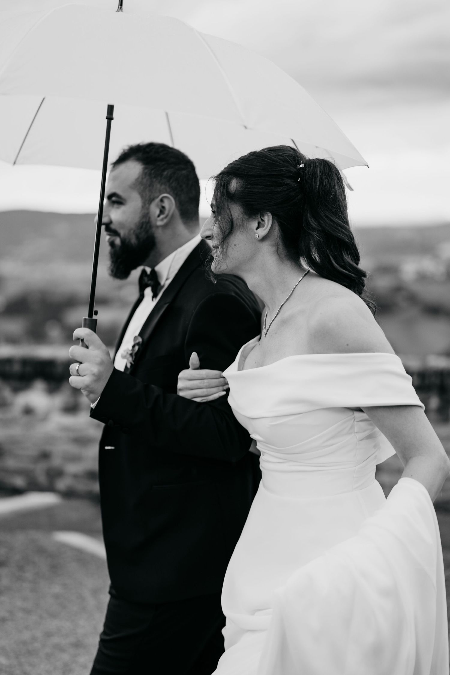 Couple sharing intimate moments under a white umbrella against a cloudy countryside backdrop.