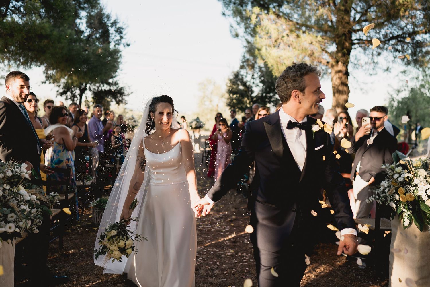Wedding couple walking down aisle while guests throw flower petals in outdoor forest setting.