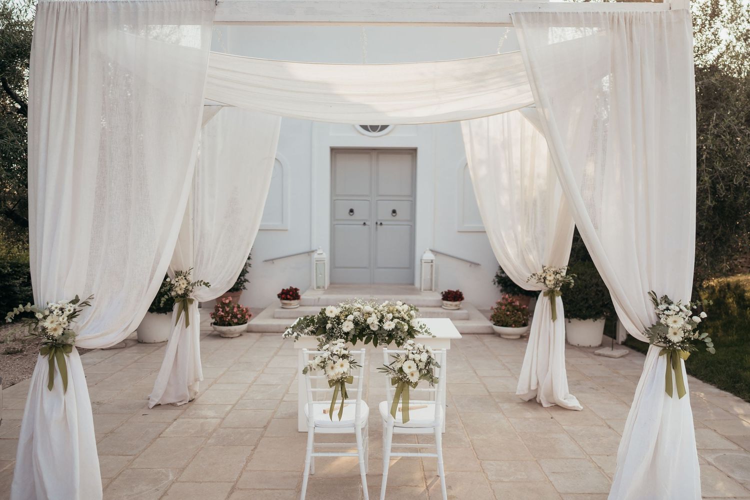 Elegant white wedding ceremony setup with flowing drapes and floral decorations against a white building.