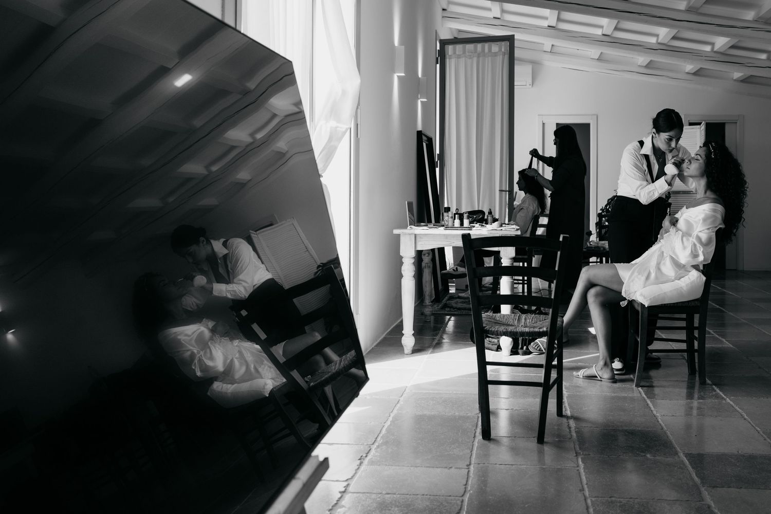 Black and white photo showing preparation scene with styling chairs and mirrors in an interior space.