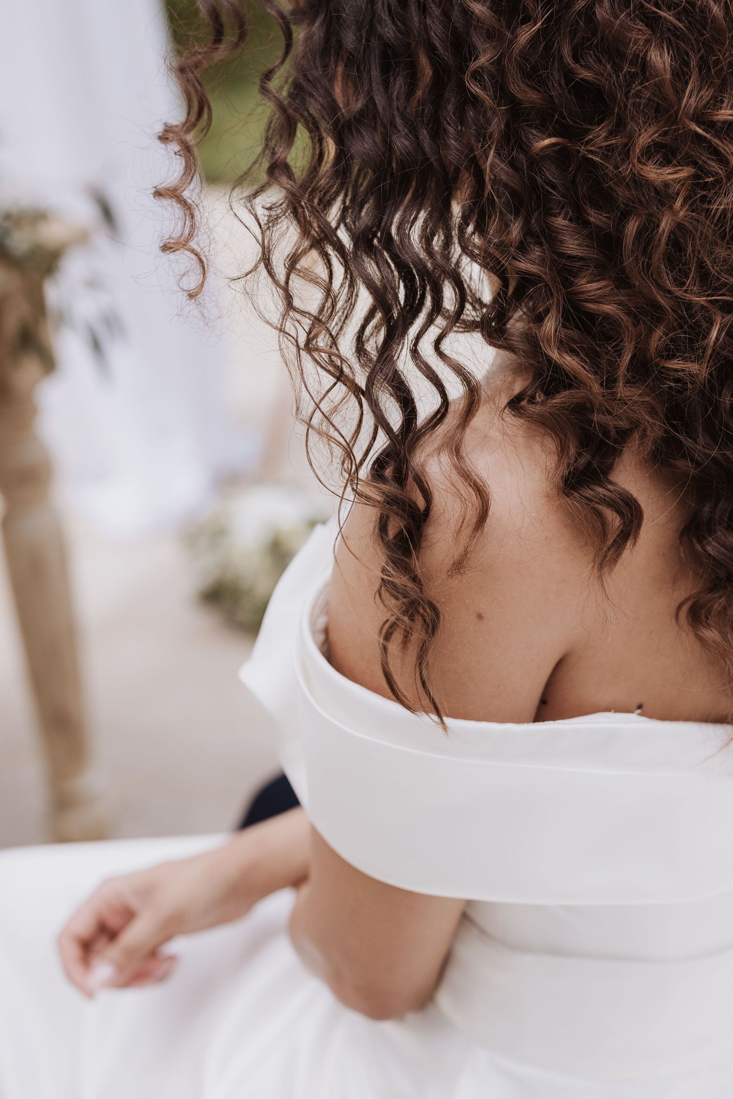 Close up detail of curly brown hair and white off-shoulder wedding dress.