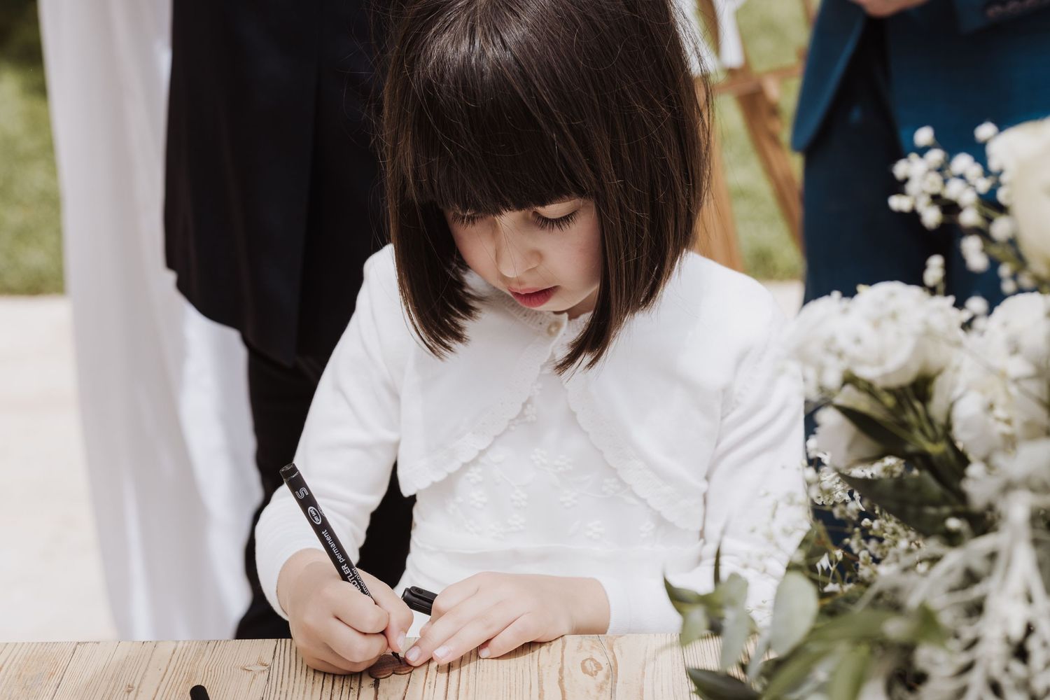 Child in white dress signing guest book at outdoor wedding.