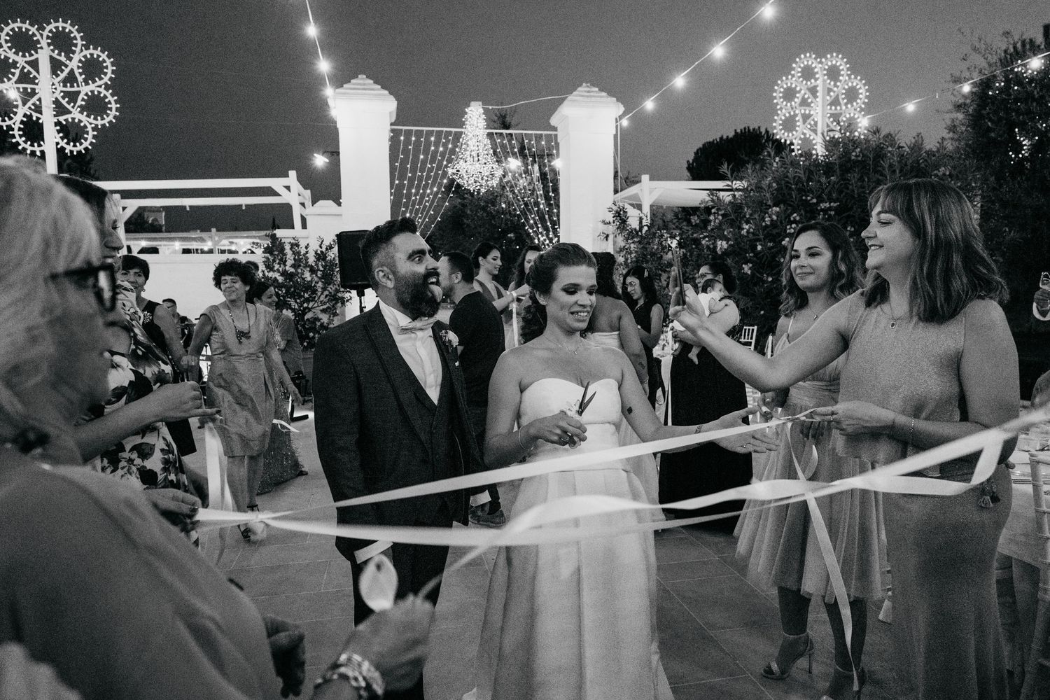 Black and white photo of wedding guests gathered around string lights at an outdoor reception.