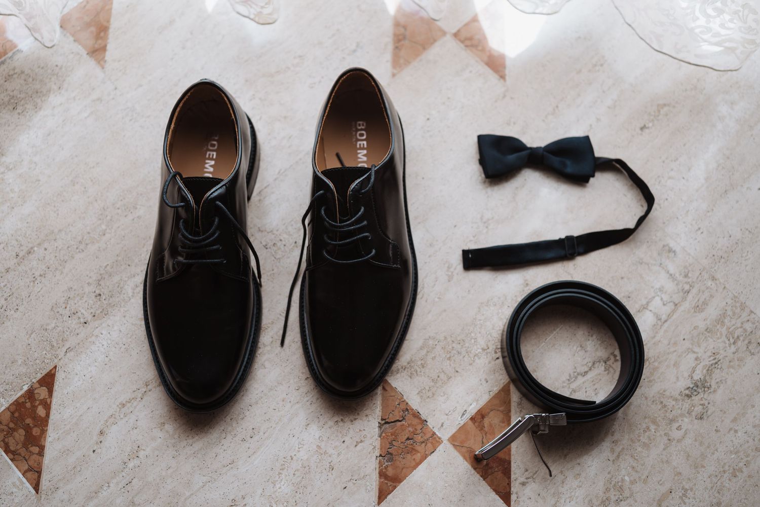 Groom's wedding accessories including black dress shoes, bow tie and belt on tile floor.