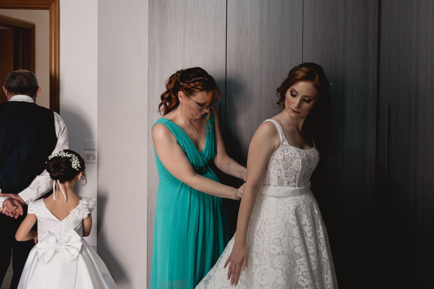Bridesmaids in turquoise dresses help adjust a white lace wedding gown during preparations.
