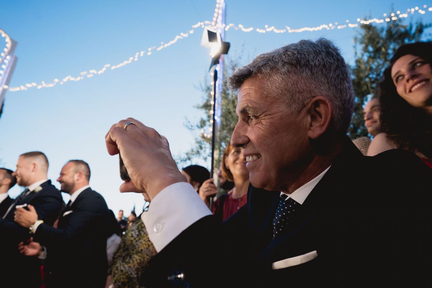 Outdoor wedding celebration with guests under string lights on sunny day.