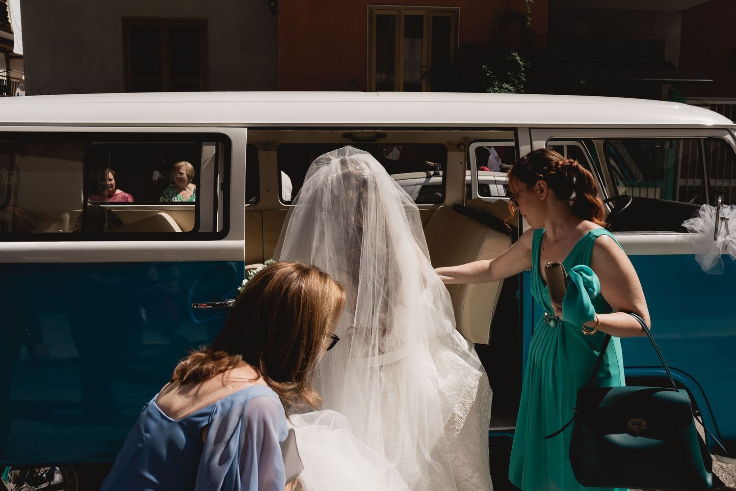 Bride in white dress and veil enters a vintage blue and white van with assistance from attendants.