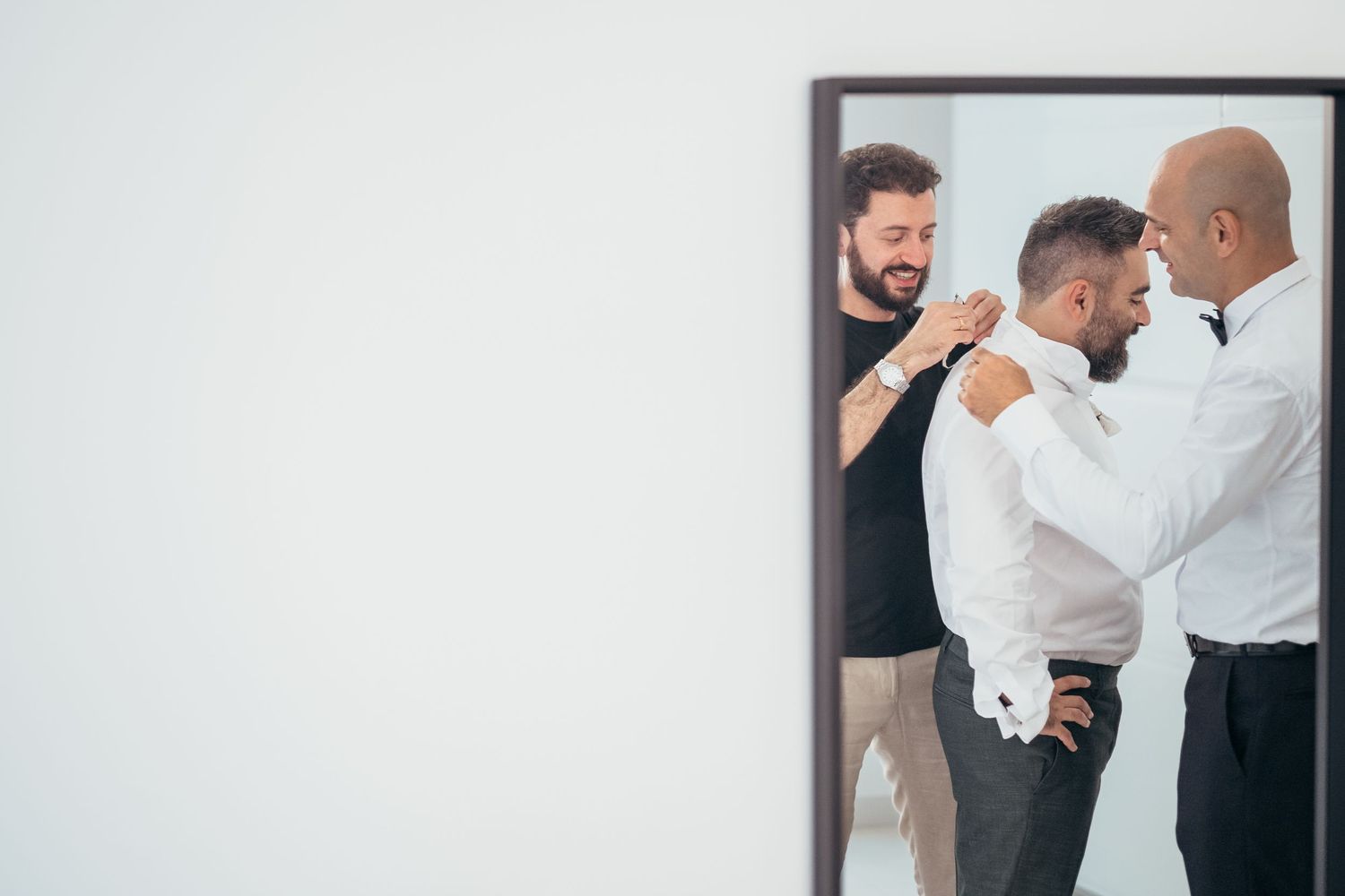 Mirror reflection shows men helping with formal attire adjustments during preparation.