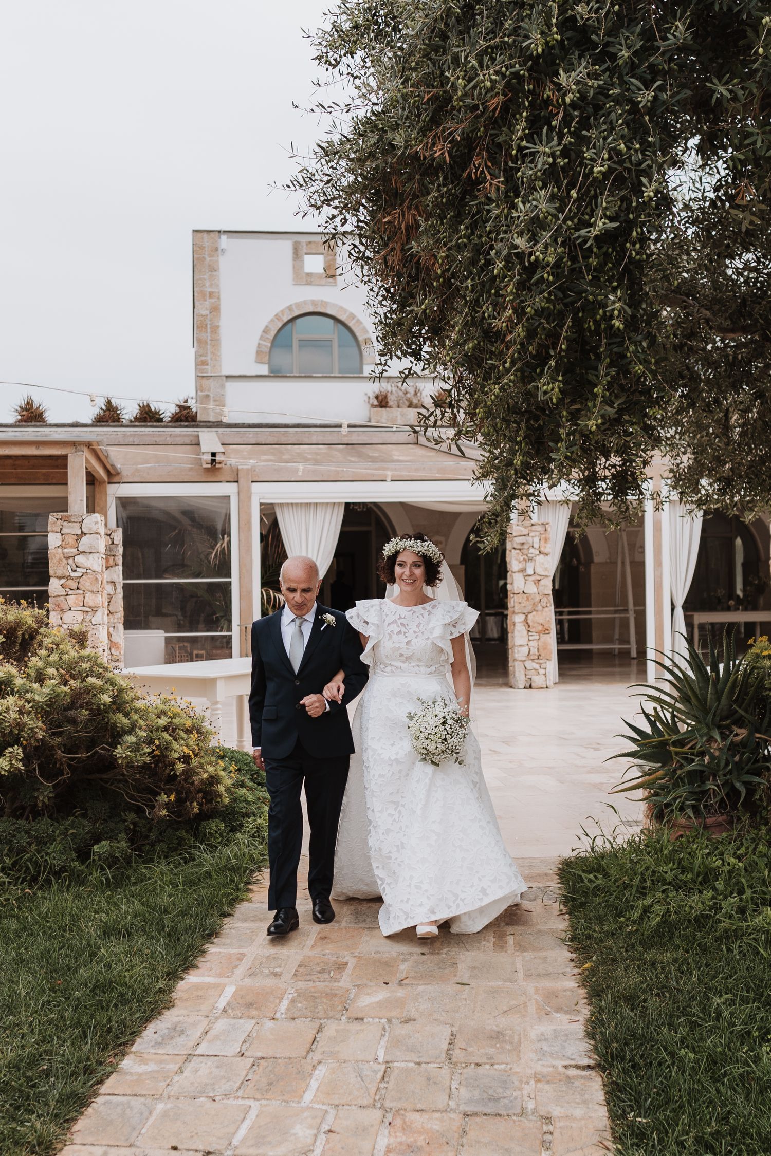 A couple walking down a stone pathway outside an elegant Mediterranean-style building.
