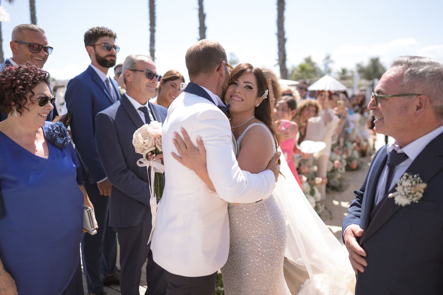 A couple shares their first kiss during an outdoor beach wedding ceremony surrounded by guests.