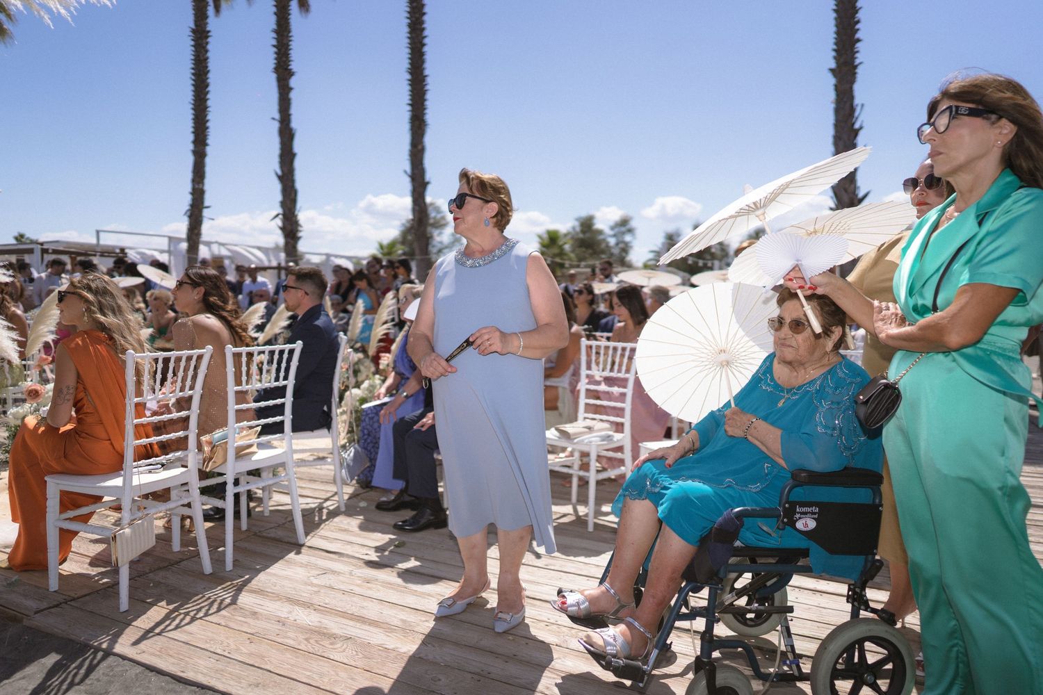 Outdoor beach wedding ceremony guests standing and seated in white chairs, woman in wheelchair holding parasol under sunny sky.