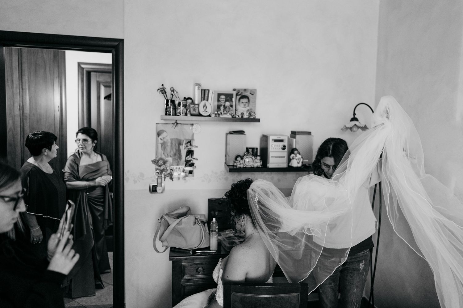 Bride getting veil fitted by stylist while family members watch in cozy room during wedding preparations.