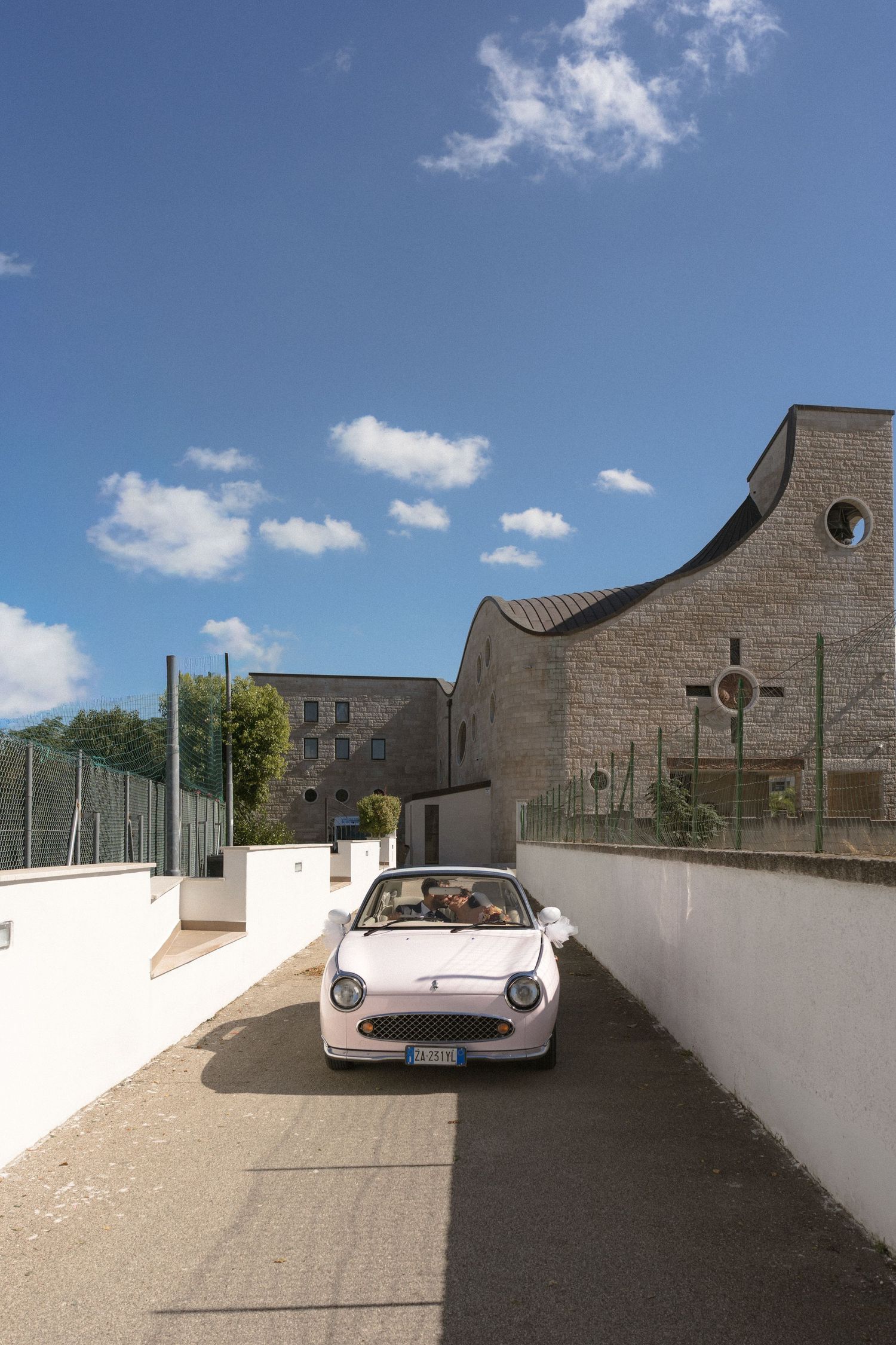 Couple rides in white vintage convertible car outside stone church on sunny wedding day.