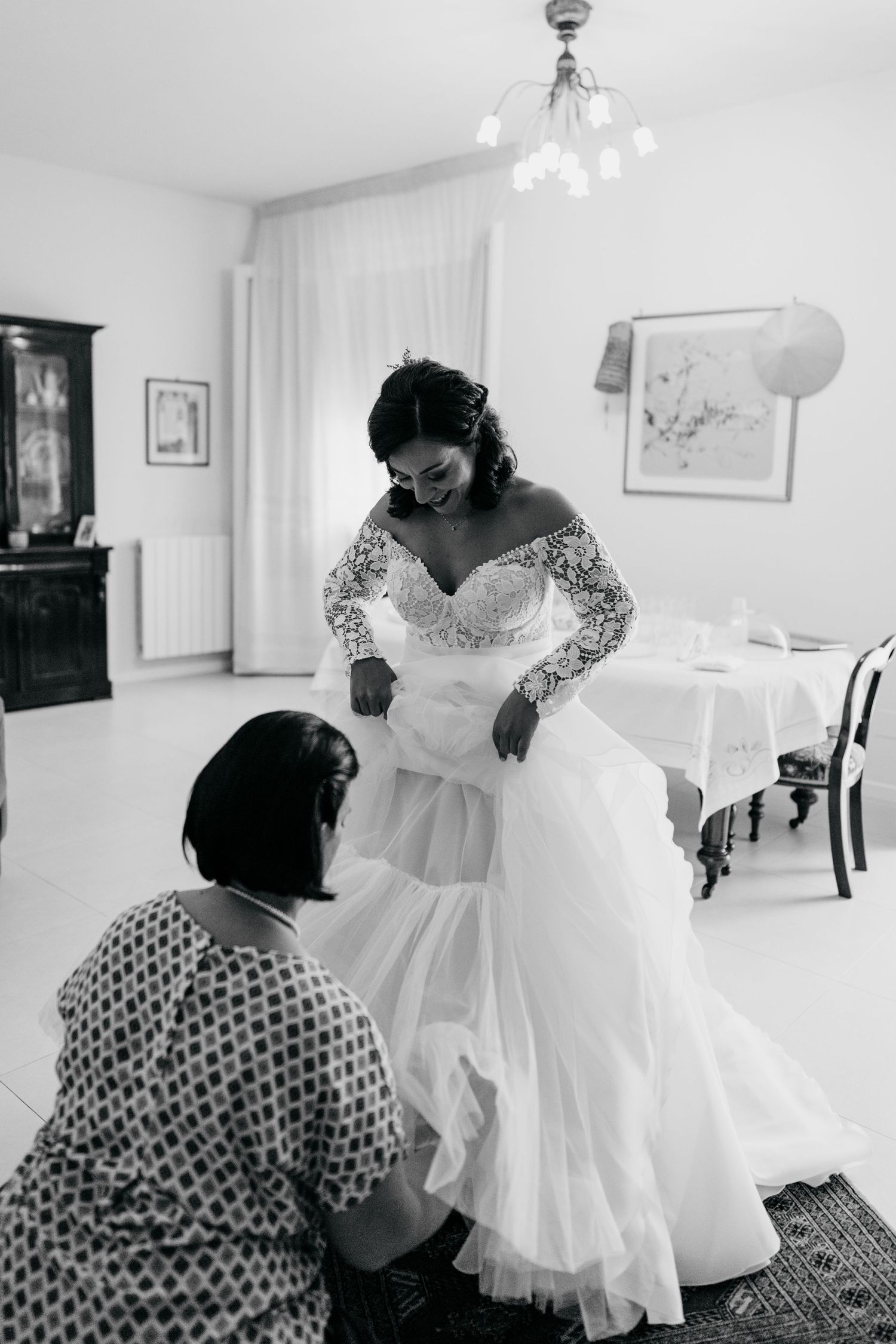 A bride in a white lace gown twirls her dress while getting ready in a bright room.