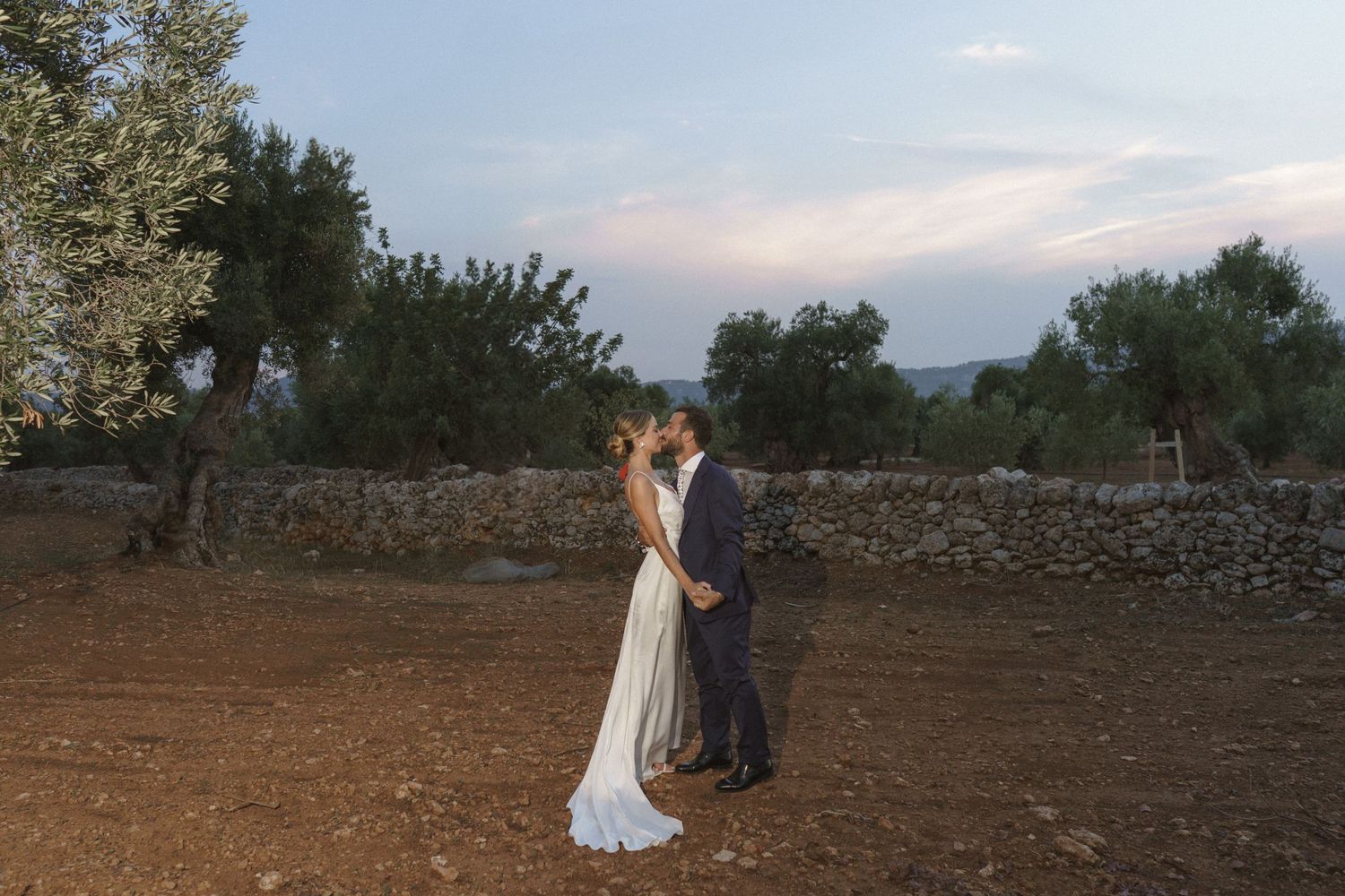 A romantic couple embracing at sunset in a rustic countryside setting with stone walls and olive trees.