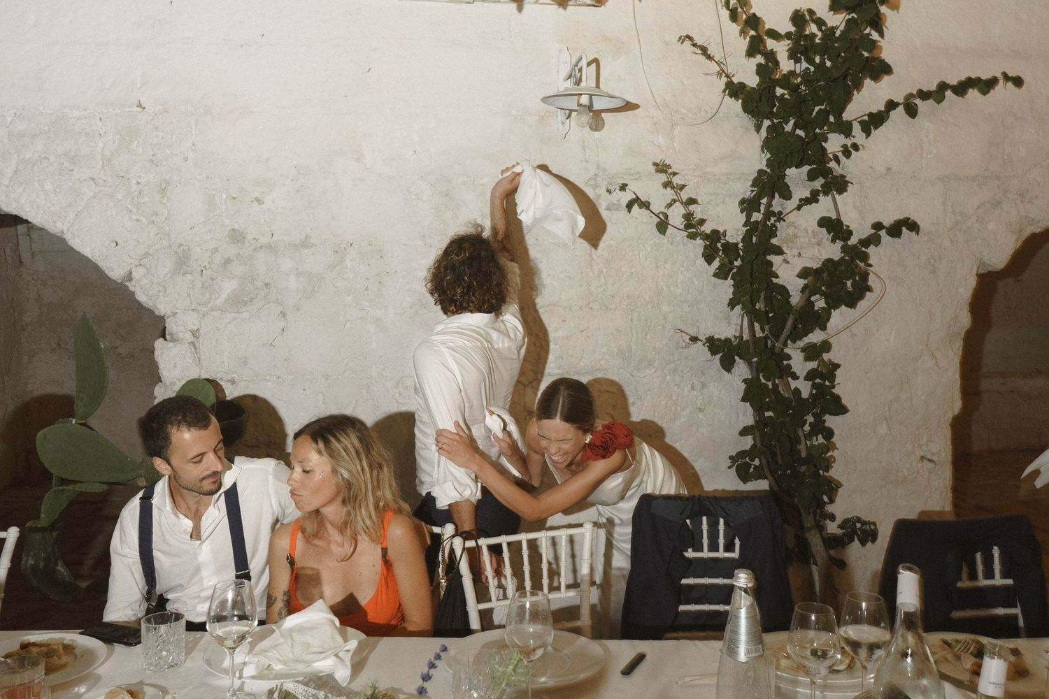 Guests enjoy a lively moment during an indoor reception as someone reaches up toward climbing vines on a white wall.