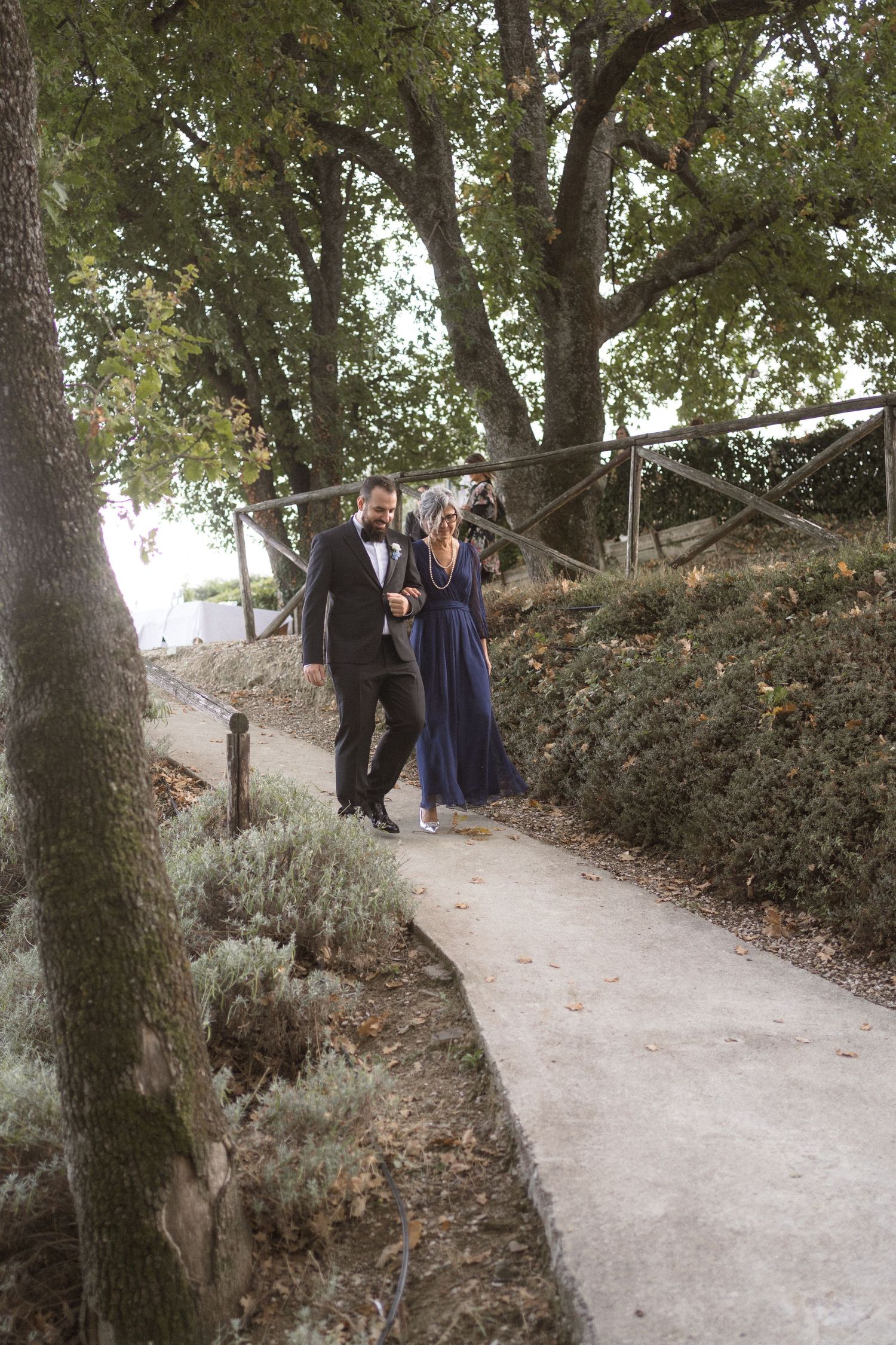 A scenic garden pathway winds through trees and foliage with people walking in formal attire.
