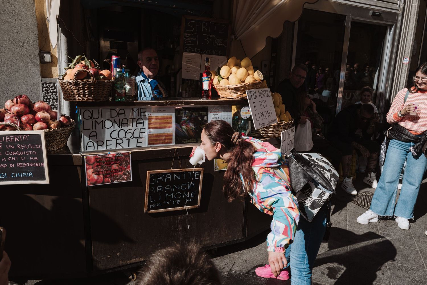 Visitors gather at a bustling market stall with chalkboard signs and merchandise displays in a dimly lit indoor setting.