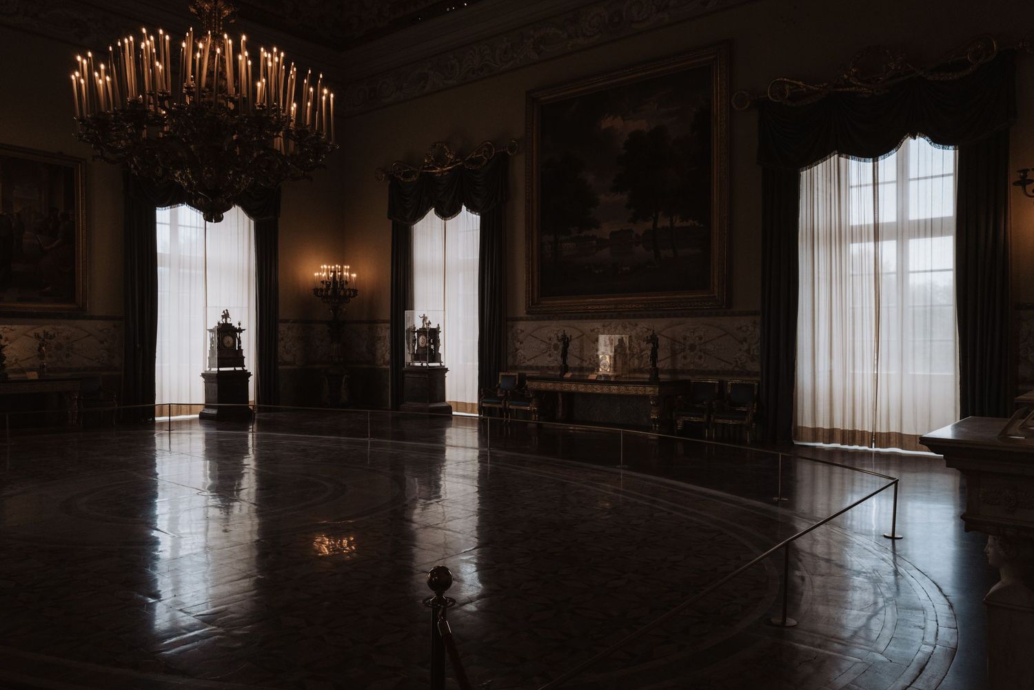 An ornate crystal chandelier illuminates a grand historic ballroom with tall windows and polished floors.