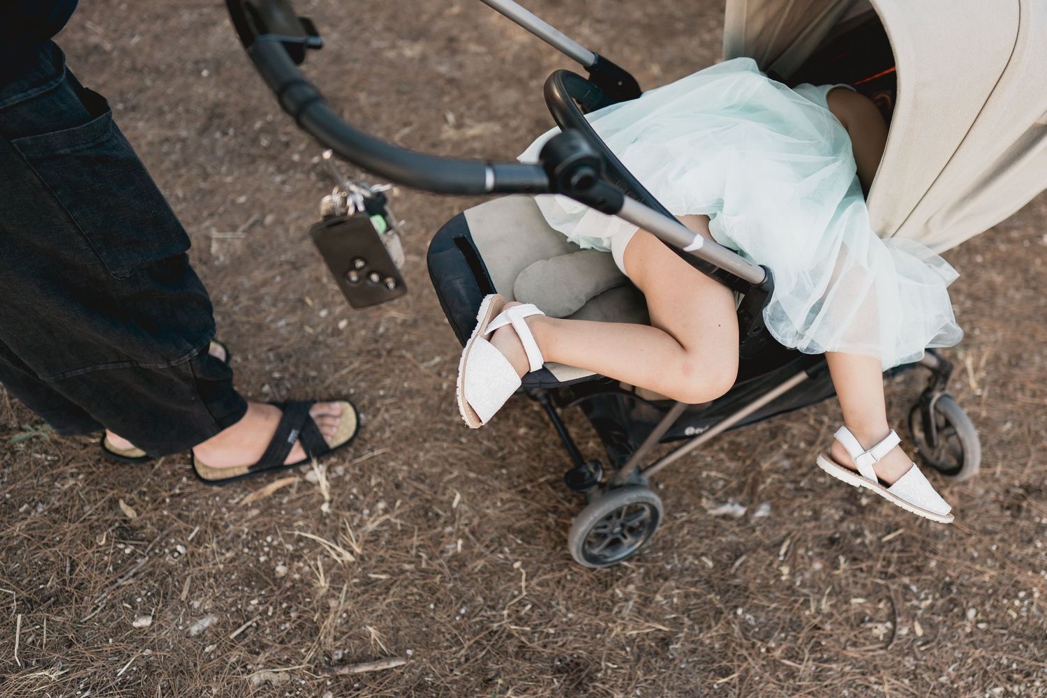 A close-up view of shoes and a stroller wheel on dirt ground.