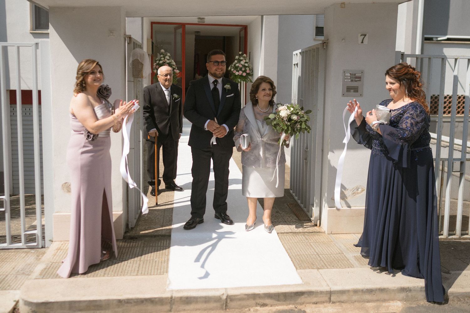 A group of people in formal attire stand in front of a white building entrance during a wedding celebration.