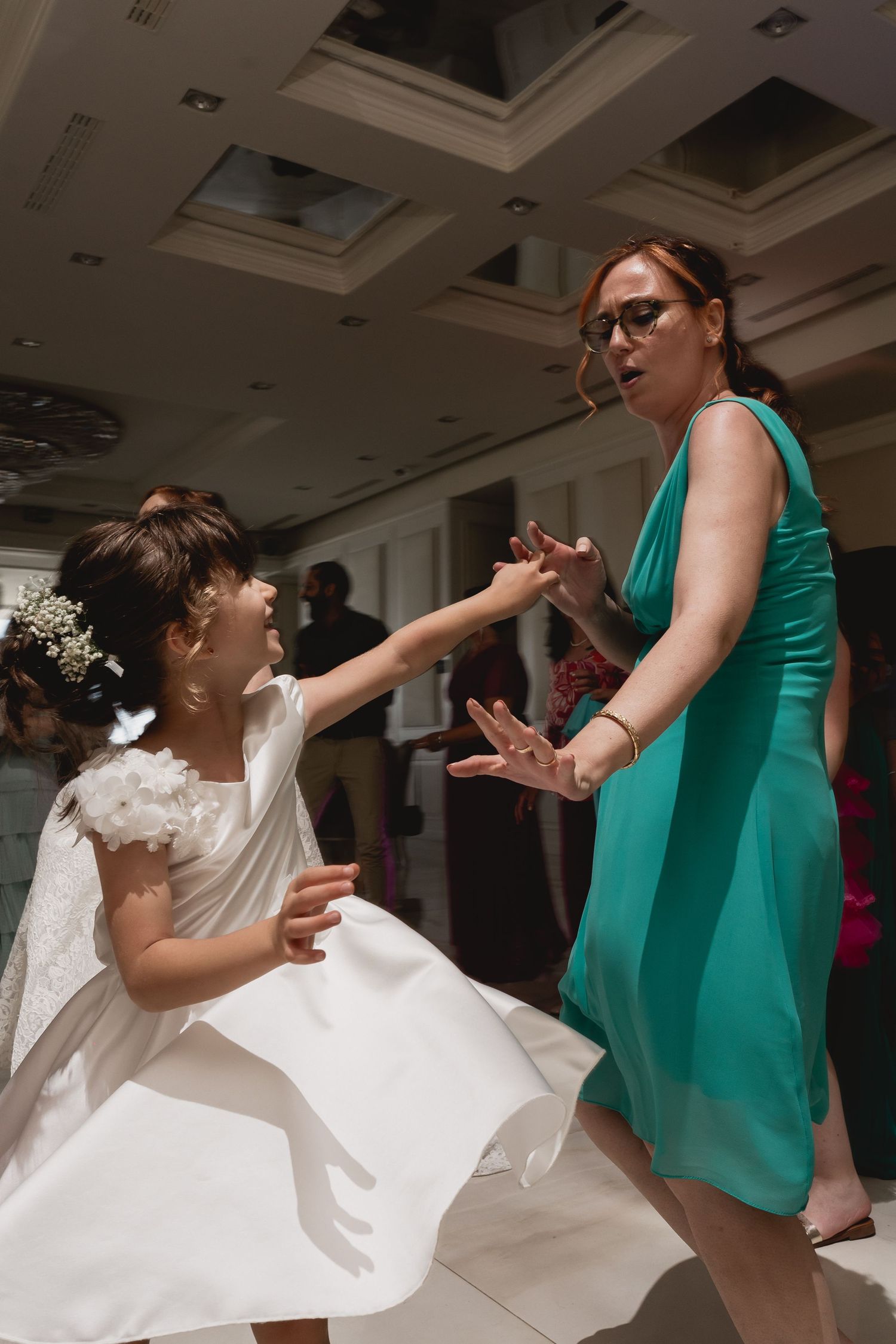 Bride in white dress dances playfully with guest in emerald green dress at reception.