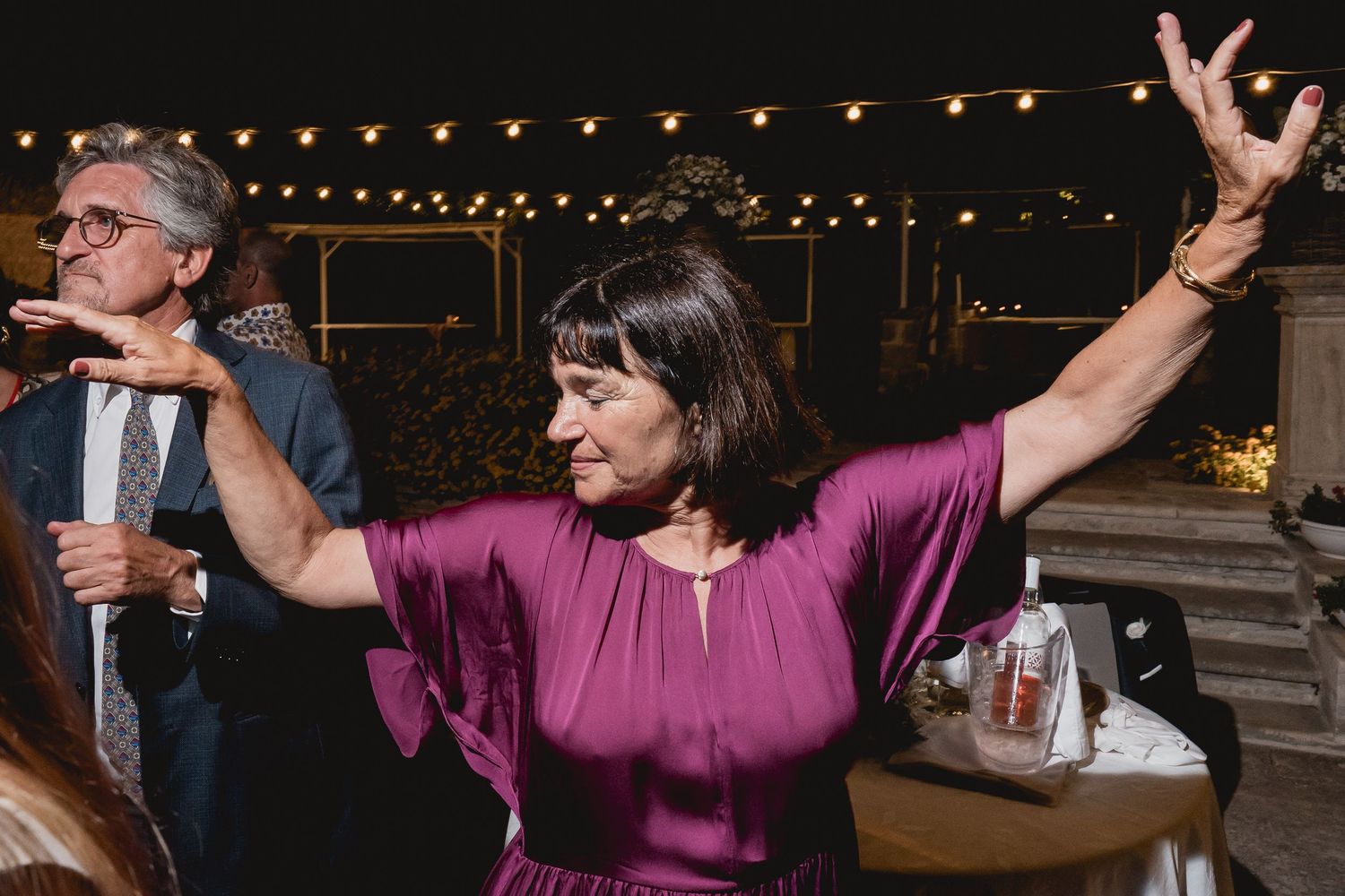 A guest in a purple dress dances enthusiastically under string lights at an outdoor reception.