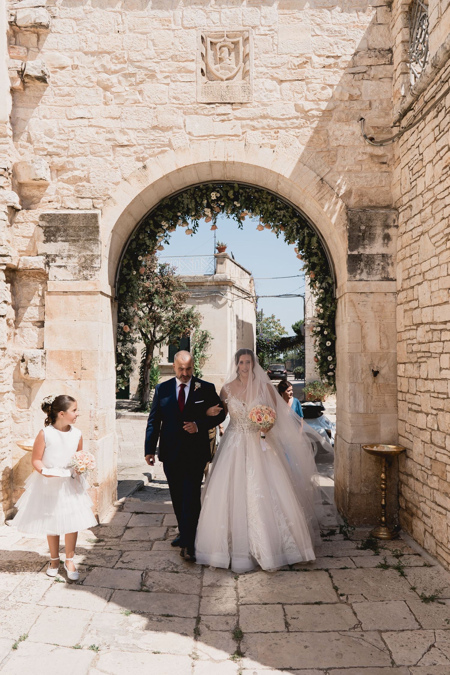 A bridal procession walking through a stone archway in traditional Italian architecture.