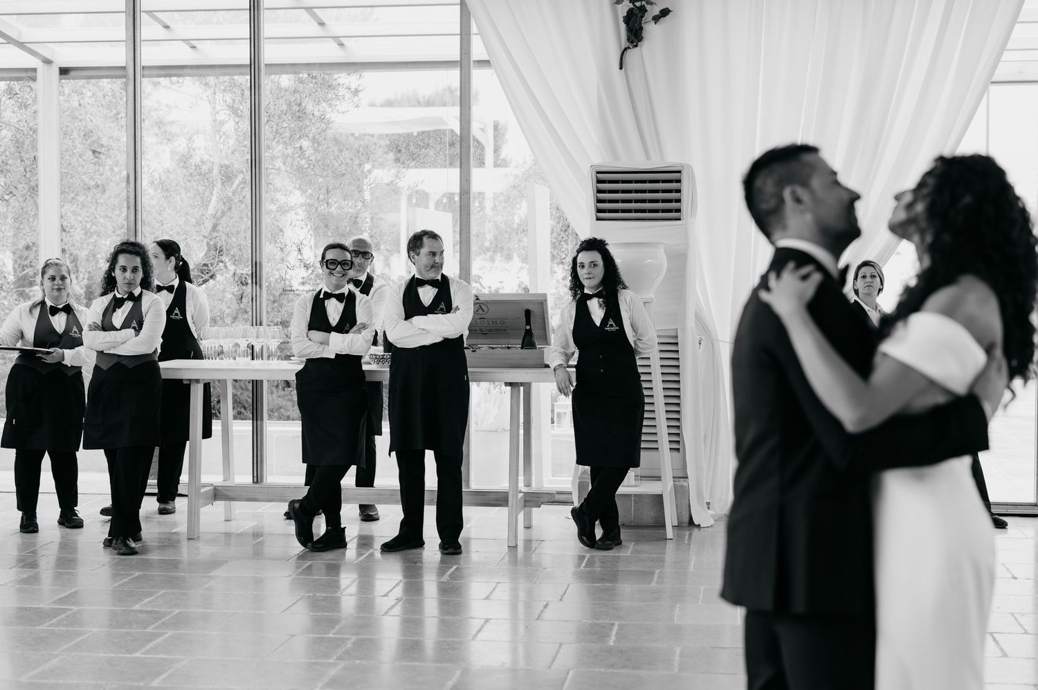 Black and white photo of wedding party members watching as couple dances at reception.