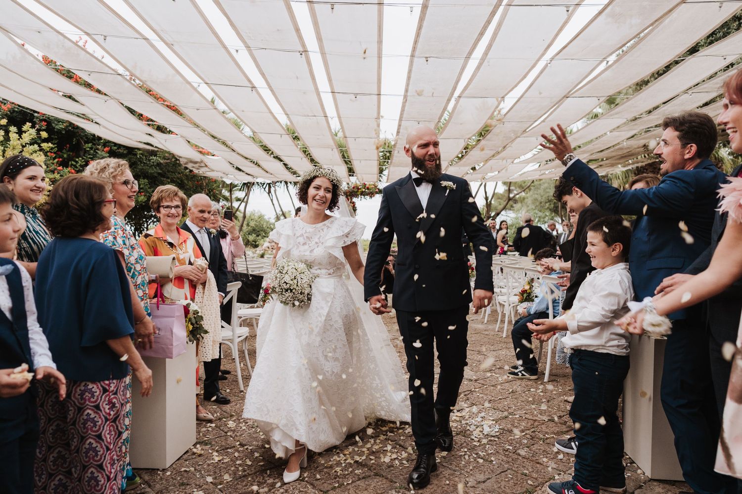 Happy couple walks down aisle while guests throw confetti at garden wedding.
